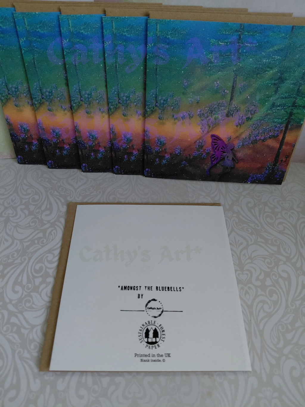 A set of artwork greeting cards with a colourful forest illustration, of a bluebell forest featuring a fairy, displayed on a patterned surface with a white card in front showing the title 'Amongst the Bluebells' by Cathy's Art*  Original artwork greeting cards by Cathy's Art*