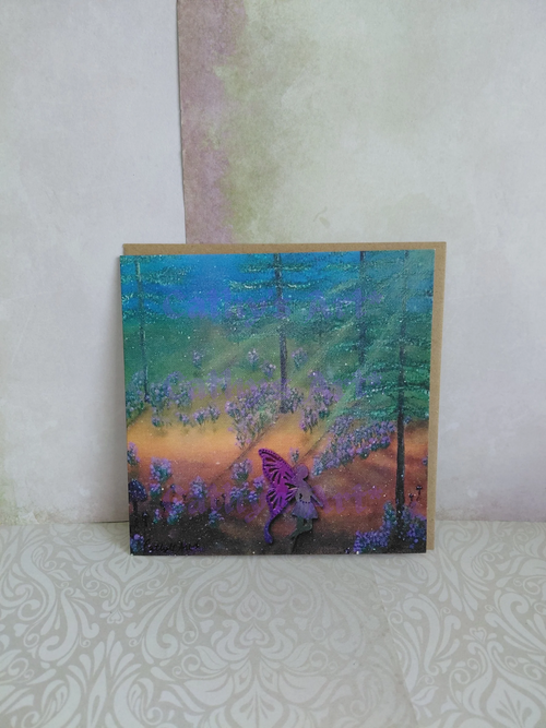 A square printed greeting card featuring a colourful forest scene with tall trees, purple bluebell flowers, and a purple fairy, displayed against a textured light wall. With a brown envelope behind the card. Original artwork printed greeting card by Cathy's Art*