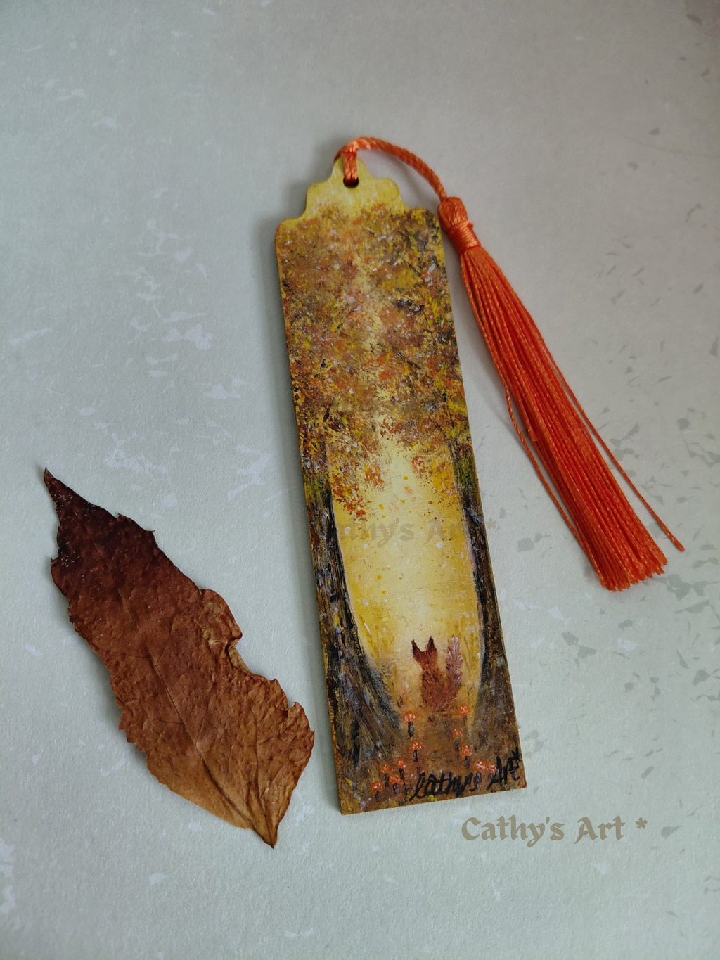 A hand painted bookmark with a orange tassel, featuring a whimsical forest scene in autumn colours, two trees, a fox sat under the trees where little orange mushrooms grow, displayed next to a dried brown leaf on a light textured background. The text 'Cathy's Art*' is visible in the lower right corner. Original hand painted bookmark by Cathy's Art*