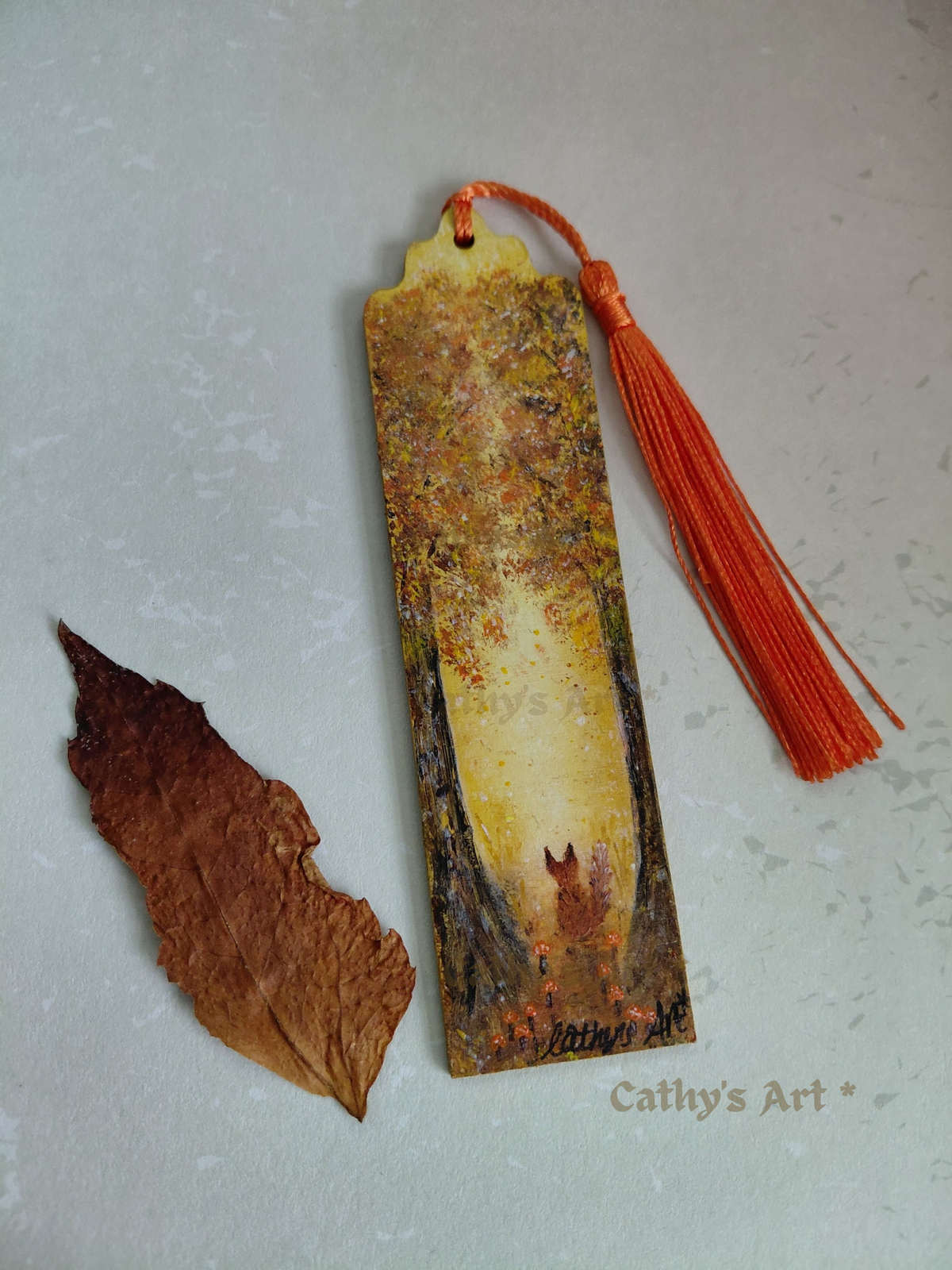 A hand painted bookmark with a orange tassel, featuring a whimsical forest scene in autumn colours, two trees, a fox sat under the trees where little orange mushrooms grow, displayed next to a dried brown leaf on a light textured background. The text 'Cathy's Art*' is visible in the lower right corner. Original hand painted bookmark by Cathy's Art*