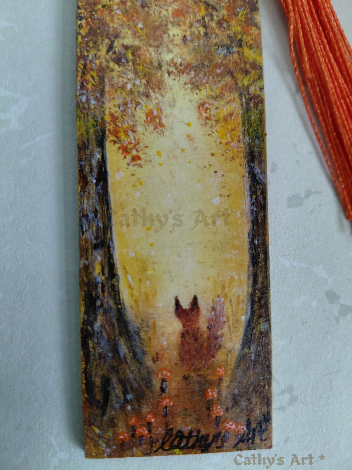 A close up photo of a hand painted autumn woodland scene bookmark featuring trees, a fox orange mushrooms, and part of an orange tassel. On a light textured background. Original hand painted bookmark by Cathy's Art*