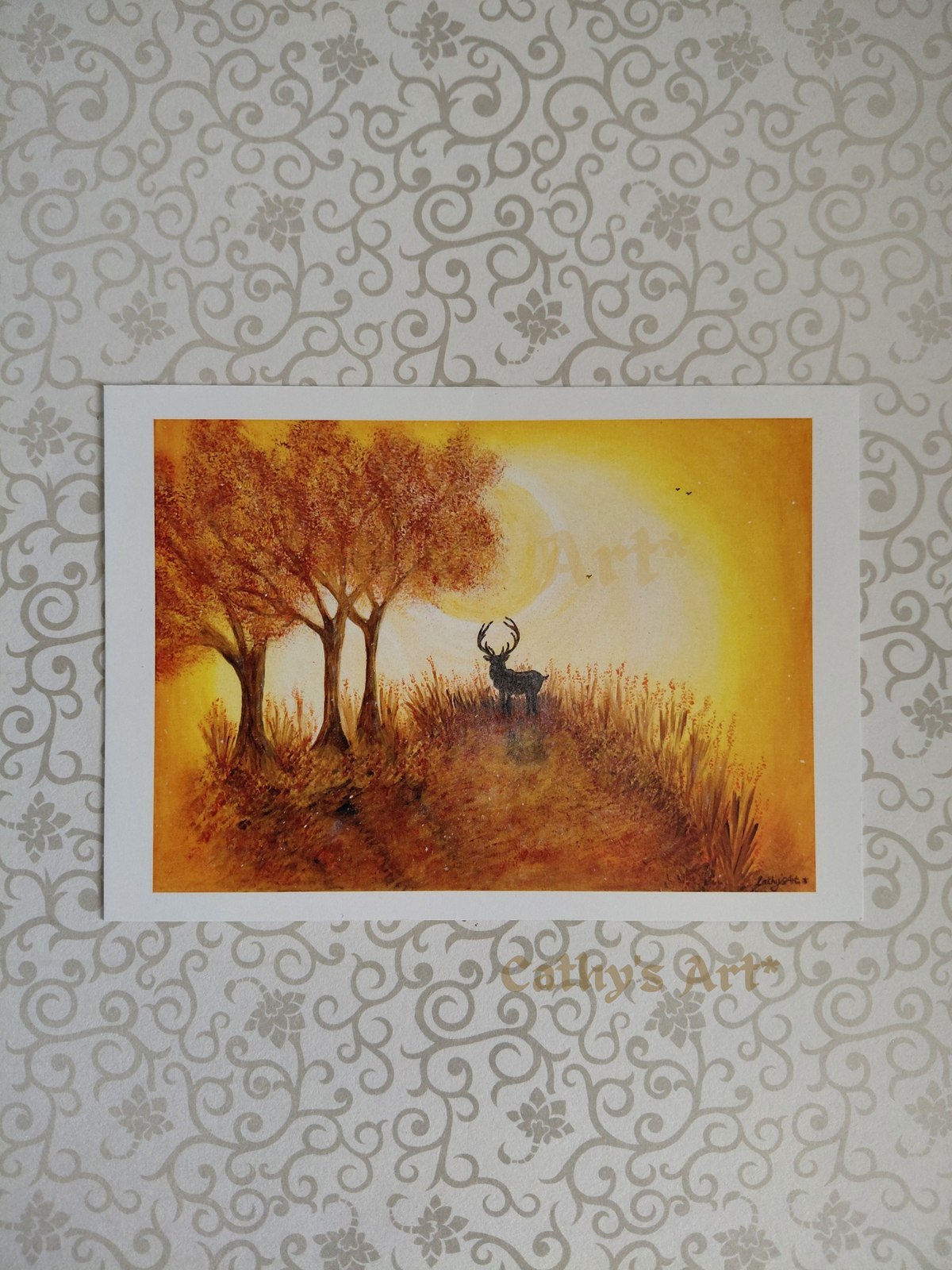 A art print featuring a painted autumn landscape with three trees, a stag, and a glowing yellow-orange sky, displayed against a patterned grey wallpaper background. The print has a white border. Artwork is by Cathy's Art* 