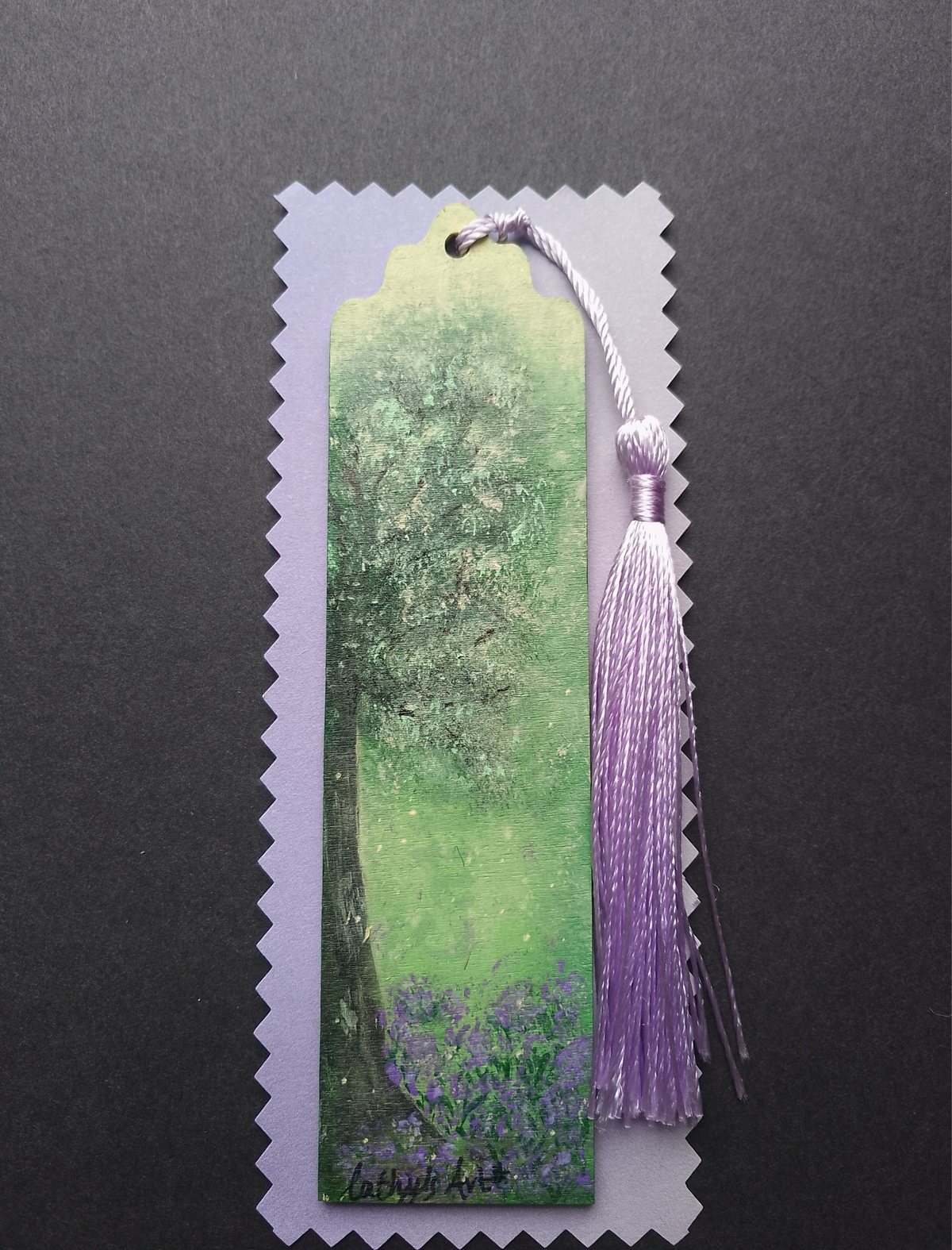 A decorative bookmark featuring a hand painted green and purple tree, purple bluebell flowers below the tree, a lilac purple tassel on lilac packaging on a black background. Original hand  bookmark by Cathy's Art*