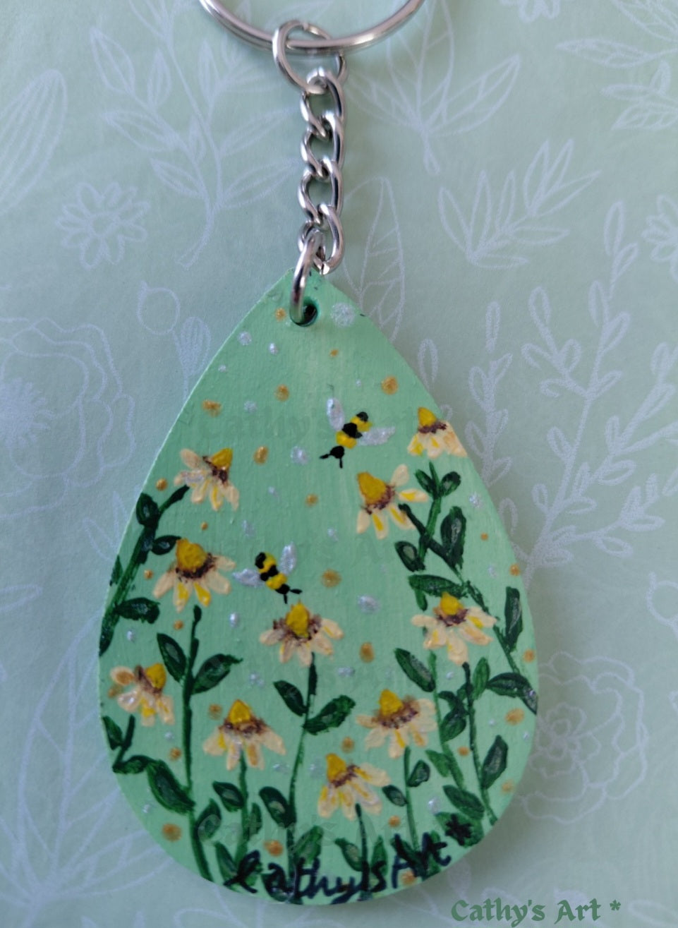 A close up teardrop-shaped wooden keychain featuring a hand painted design of yellow daisy flowers, green leaves, and bumblebees on a mint green background, with a silver metal split ring. Original acrylic keyring by Cathy's Art*