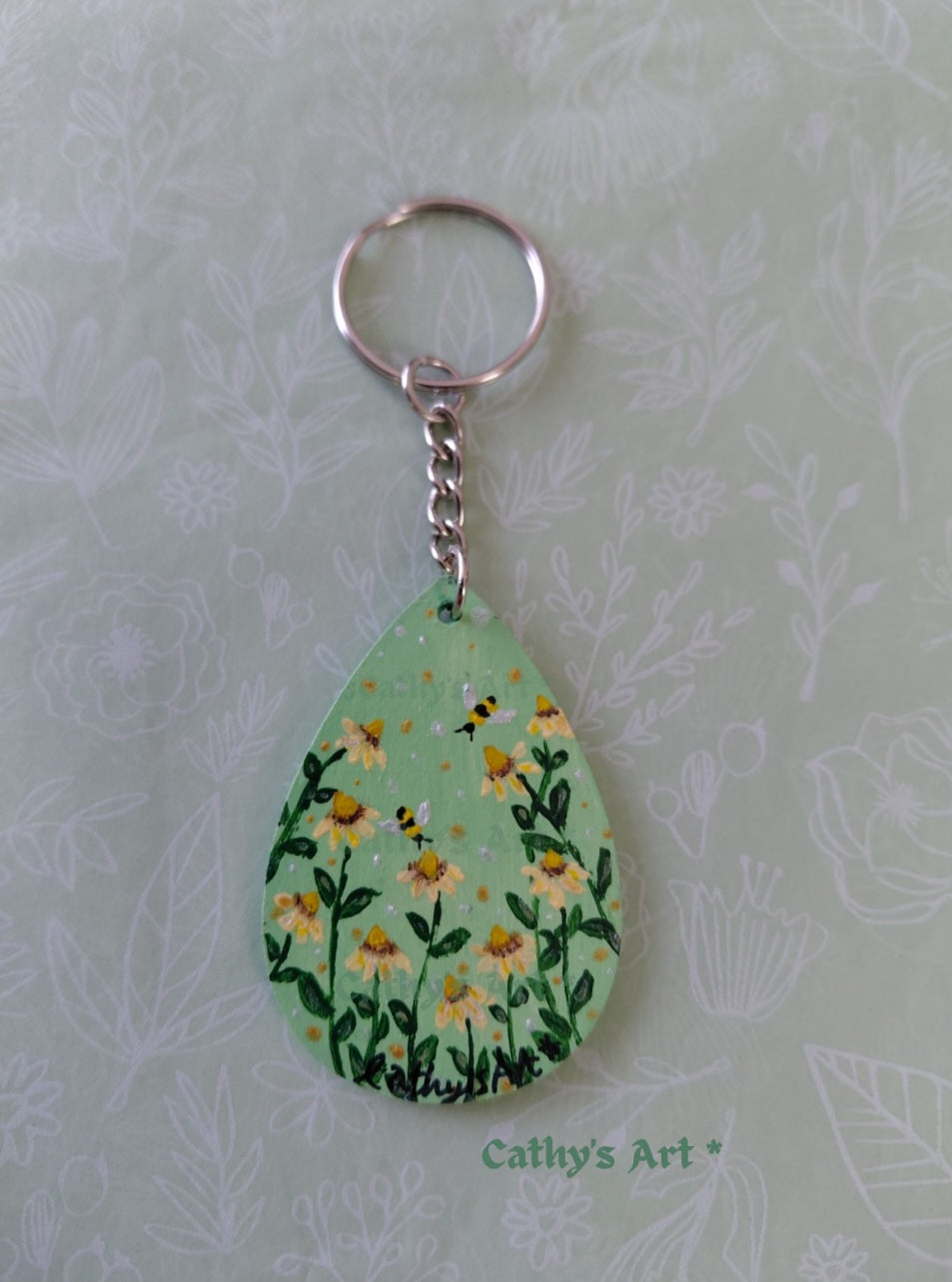 A teardrop-shaped wooden keychain featuring a hand painted green background with yellow daisy flowers, two bees, attached to a silver metal key ring and chain. The keychain is displayed on a light green floral-patterned background with the text 'Cathy's Art' visible at the bottom. Original acrylic painted keyring by Cathy's Art*