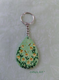 A teardrop-shaped wooden keychain featuring a hand painted green background with yellow daisy flowers, two bees, attached to a silver metal key ring and chain. The keychain is displayed on a light green floral-patterned background with the text 'Cathy's Art' visible at the bottom. Original acrylic painted keyring by Cathy's Art*