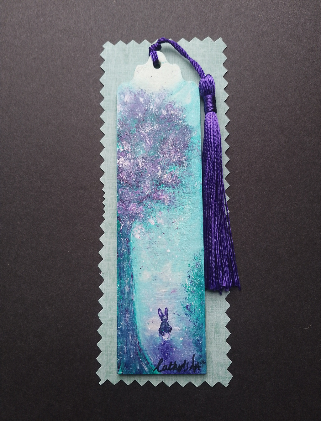 A hand painted whimsical bookmark with a purple tassel, featuring an artistic painting of a moonlit tree, in shades of turquoise, purple, and white, with a small rabbit illustration near the bottom. Photographed on light packaging, Signed "Cathy's Art*" at the bottom. Original hand painted bookmark by Cathy's Art*