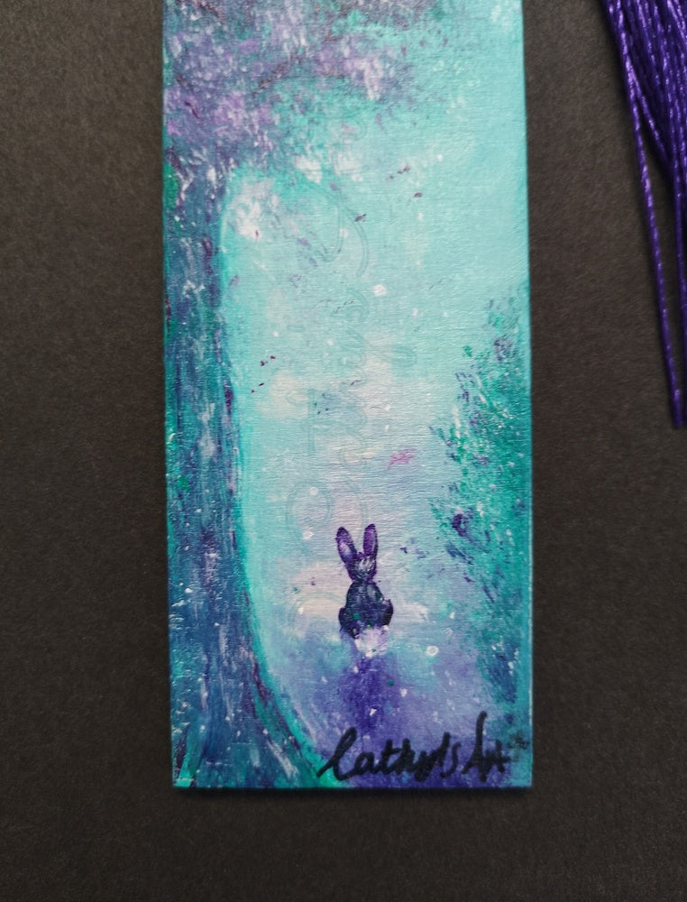 A close up of a hand painted bookmark with a whimsical design, in turquoise, white and purple colours, a little rabbit sits under the purple tree. On a black back ground. original hand painted bookmark by Cathy's Art*