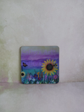 A square decorative coaster with rounded corners featuring a colourful painted landscape of sunflowers, wildflowers, a butterfly, a bee and a purple sky, displayed on a textured light beige surface. Original artwork printed coaster by Cathy's Art*
