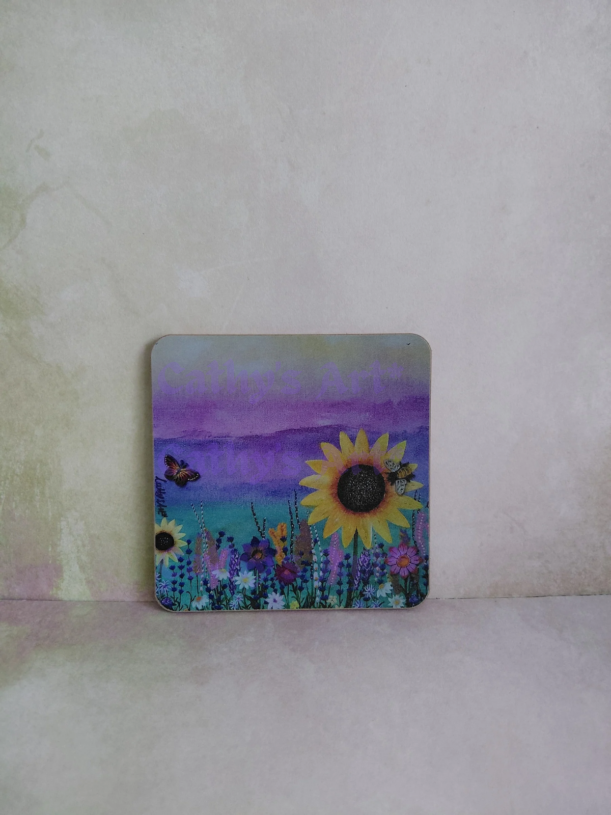 A square decorative coaster with rounded corners featuring a colourful painted landscape of sunflowers, wildflowers, a butterfly, a bee and a purple sky, displayed on a textured light beige surface. Original artwork printed coaster by Cathy's Art*