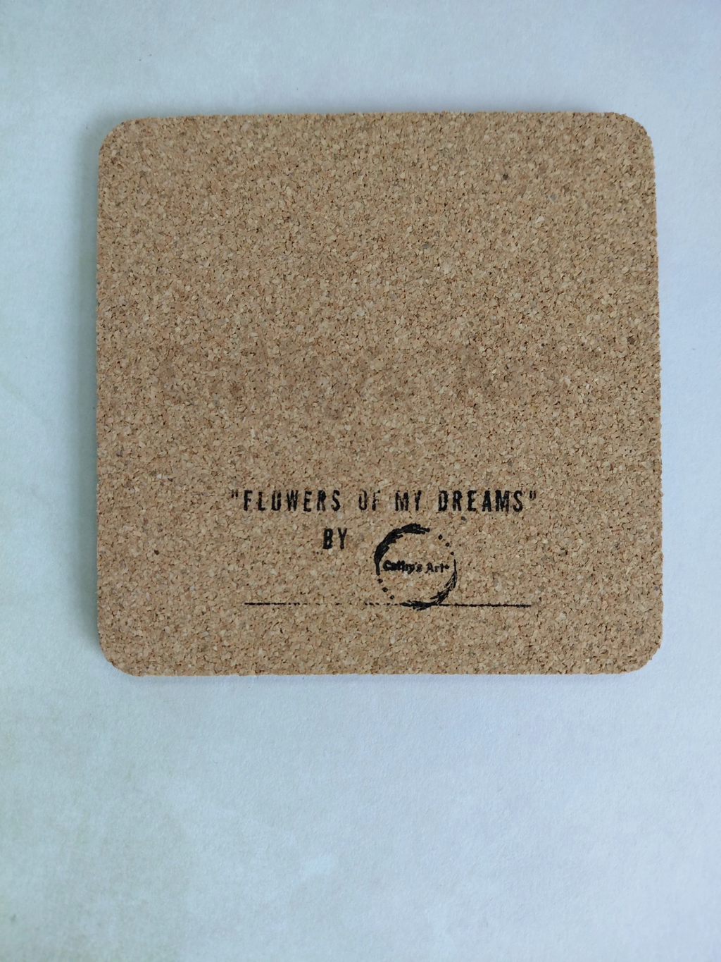 A square cork coaster with rounded corners, featuring the printed text 'FLOWERS OF MY DREAMS' BY Cathy's Art* in black ink, displayed on a light  background. Original artwork printed coaster by Cathy's Art*