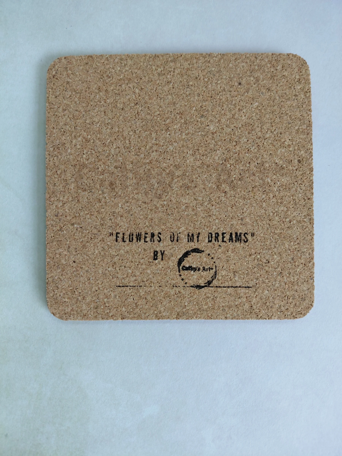 A square cork coaster with rounded corners, featuring the printed text 'FLOWERS OF MY DREAMS' BY Cathy's Art* in black ink, displayed on a light  background. Original artwork printed coaster by Cathy's Art*