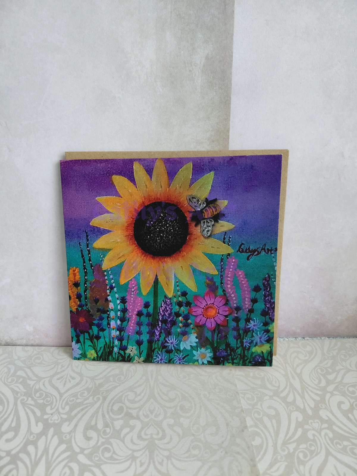 A square printed artwork greeting card featuring a colourful  floral scene with a sunflower, a bee, and wildflowers on a purple and teal green background, standing on a patterned surface against a textured wall. Original artwork greeting card by Cathy's Art*