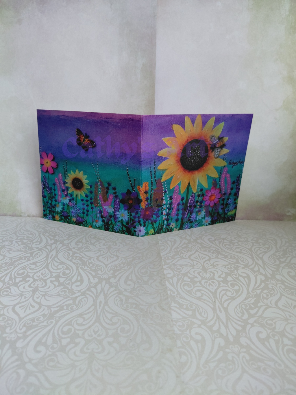 A folded greeting card featuring a colourful wildflower meadow with a large yellow sunflower, smaller flowers, butterfly and a bee, and a purple-to-teal green gradient sky, displayed against a textured light beige background. original printed artwork greeting card by Cathy's Art*