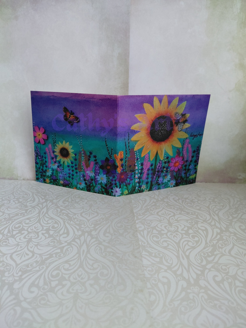 A folded greeting card featuring a colourful wildflower meadow with a large yellow sunflower, smaller flowers, butterfly and a bee, and a purple-to-teal green gradient sky, displayed against a textured light beige background. original printed artwork greeting card by Cathy's Art*