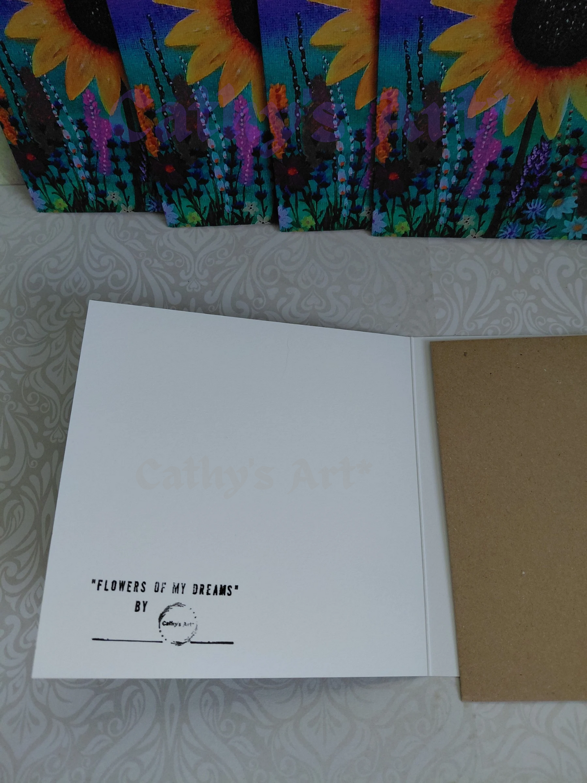 An open greeting card set on a patterned surface, showing a white page with the text 'FLOWERS OF MY DREAMS' by Cathy's Art* small circular logo, alongside a brown envelope. Sunflower floral greeting cards are  visible along the top edge. Original artwork greeting cards by Cathy's art