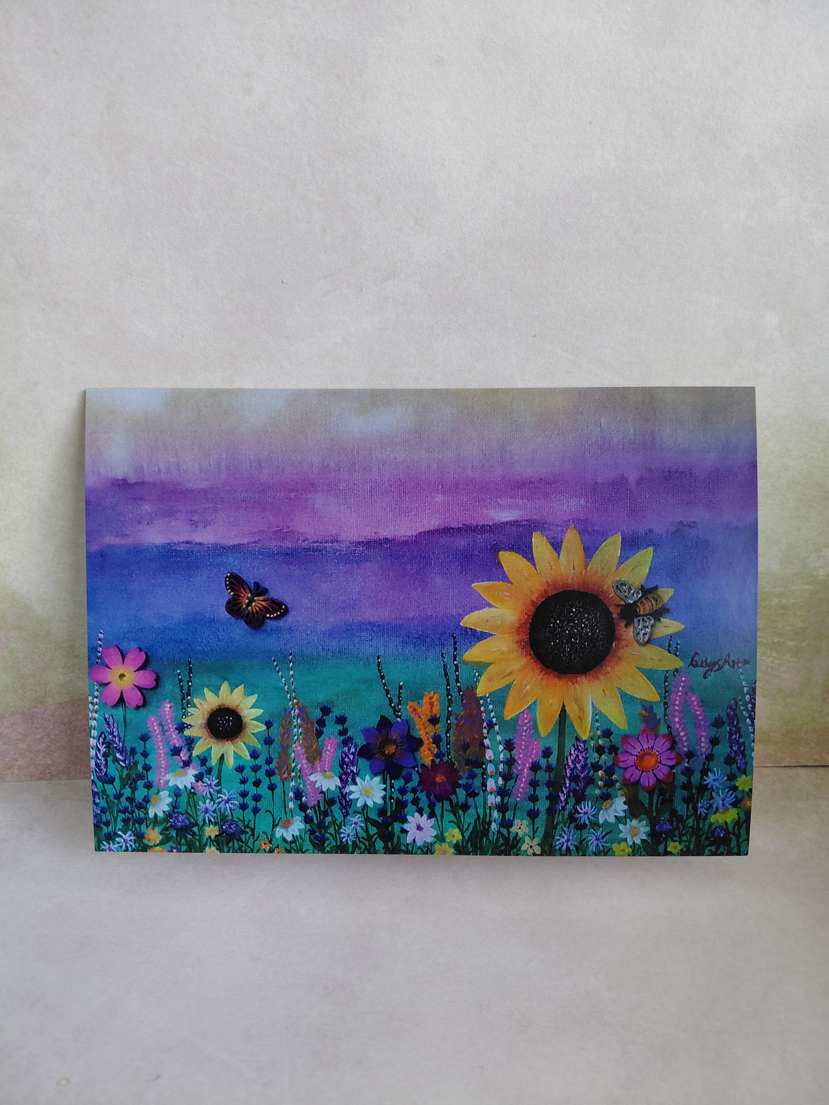 A printed tent style greeting card with a vibrant field of wildflowers with prominent sunflowers, a butterfly, and a bee against a purple sky, displayed on a textured light wall. A printed greeting card of an original painting by Cathy's art*
