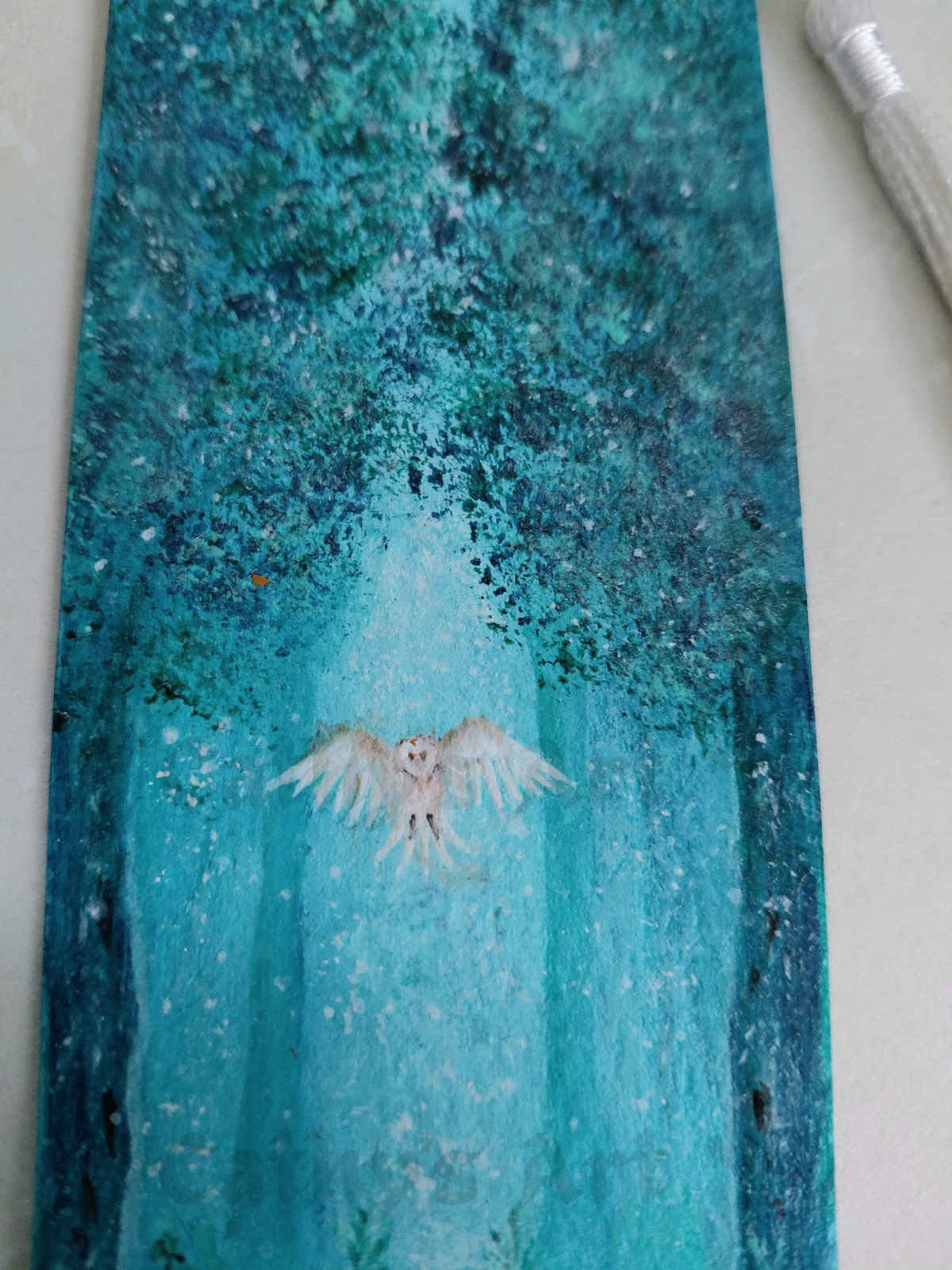 A close up shot of a hand painted bookmark featuring a painted forest scene in shades of teal, turquoise, and blue, with a white owl flying through the centre of the trees. Original acrylic painting on watercolour paper bookmark by Cathy's Art*