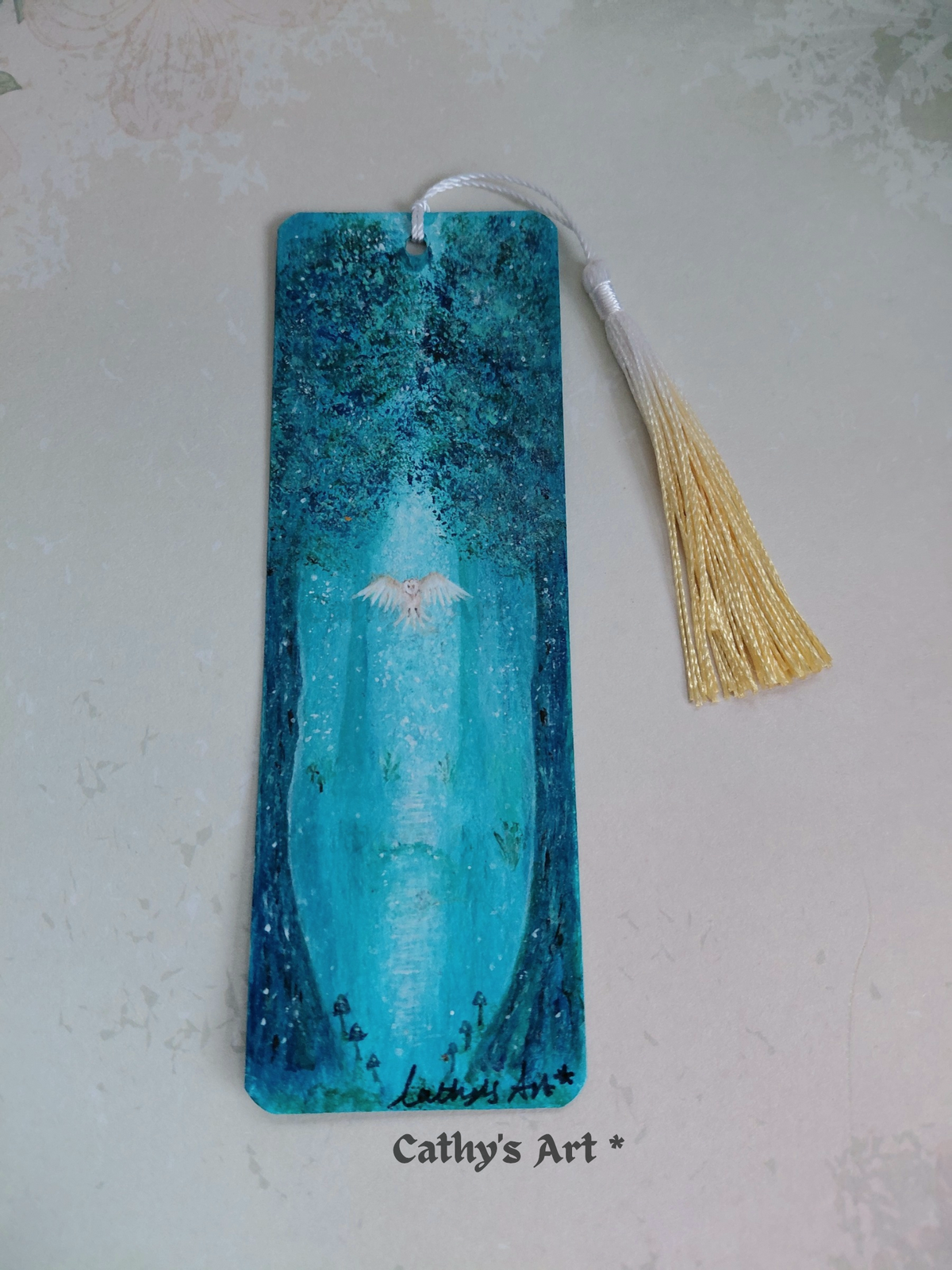 A rectangular painted bookmark with a white and yellow gold tassel, featuring a blue-toned forest scene with a white owl, two trees, tiny mushrooms and the text 'Cathy's Art *' printed near the bottom, shown on a light textured background. Original acrylic painting on watercolour paper bookmark by Cathy's Art*