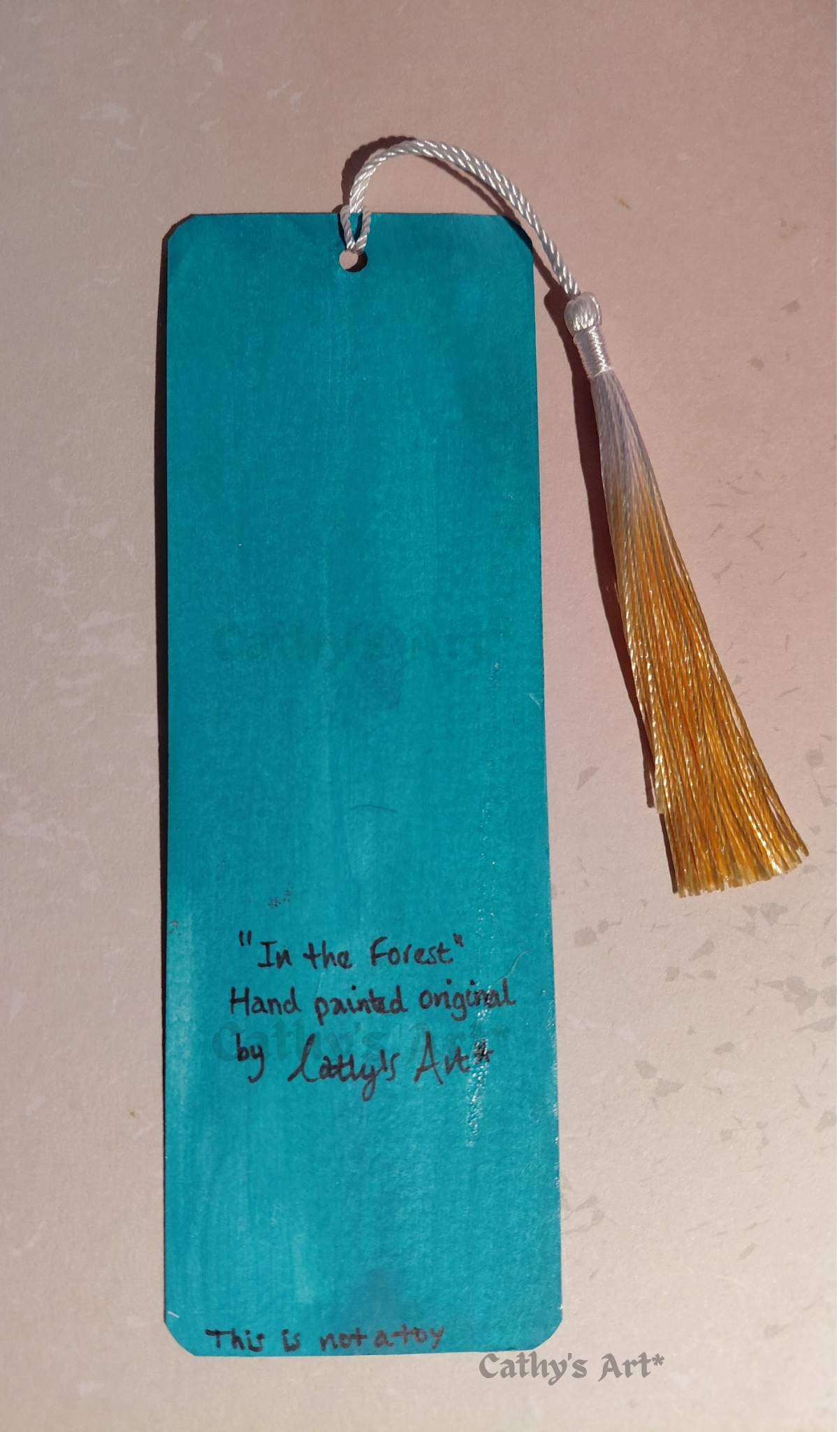 A turquoise painted bookmark with a white and yellow gold tassel, featuring hand-painted text that reads 'In the Forest' 'by Cathy's Art'. On a light coloured background. Original acrylic painting on bookmark by Cathy's art*