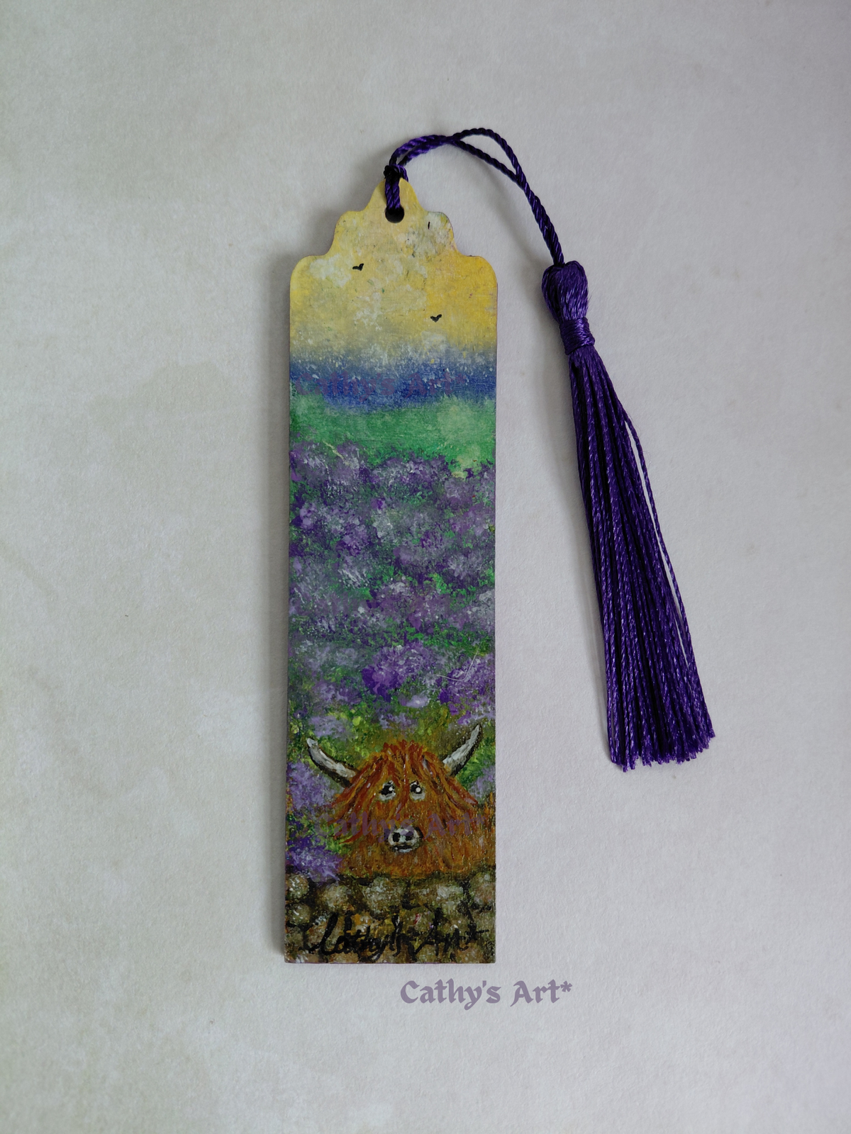 A hand-painted wooden bookmark featuring a Highland cow in a purple lavender field under a yellow and blue sky, complete with a purple tassel, displayed on a light textured background. By Cathy's Art*