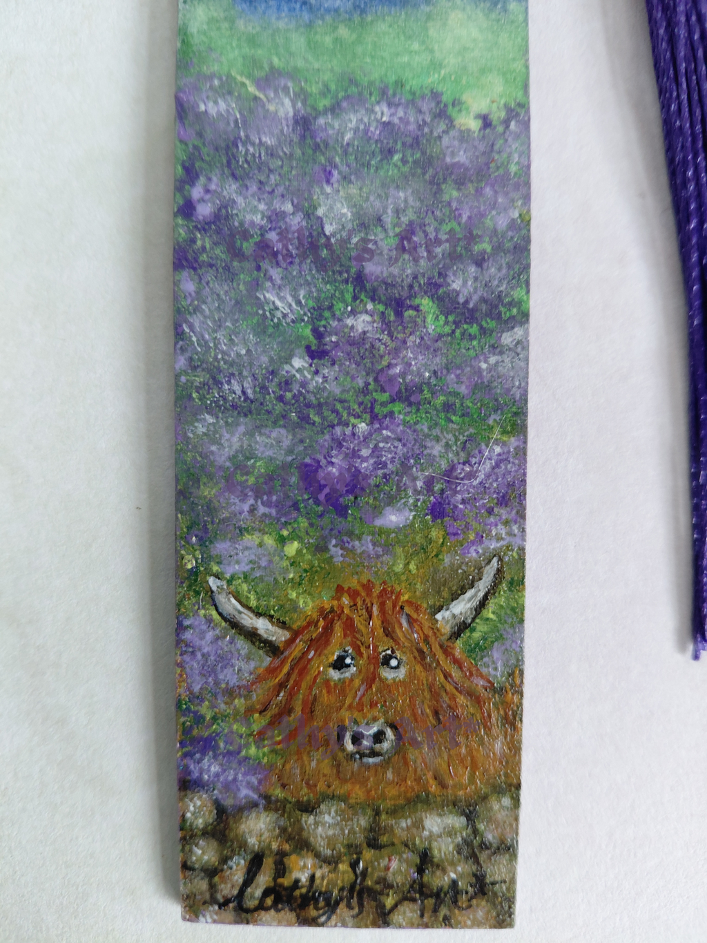 A close up view of a hand painted bookmark featuring Highland cow in front of purple heather. The cow is behind a wall. Displayed against a light background with part of a purple tassel visible at the right edge. By Cathy's Art*