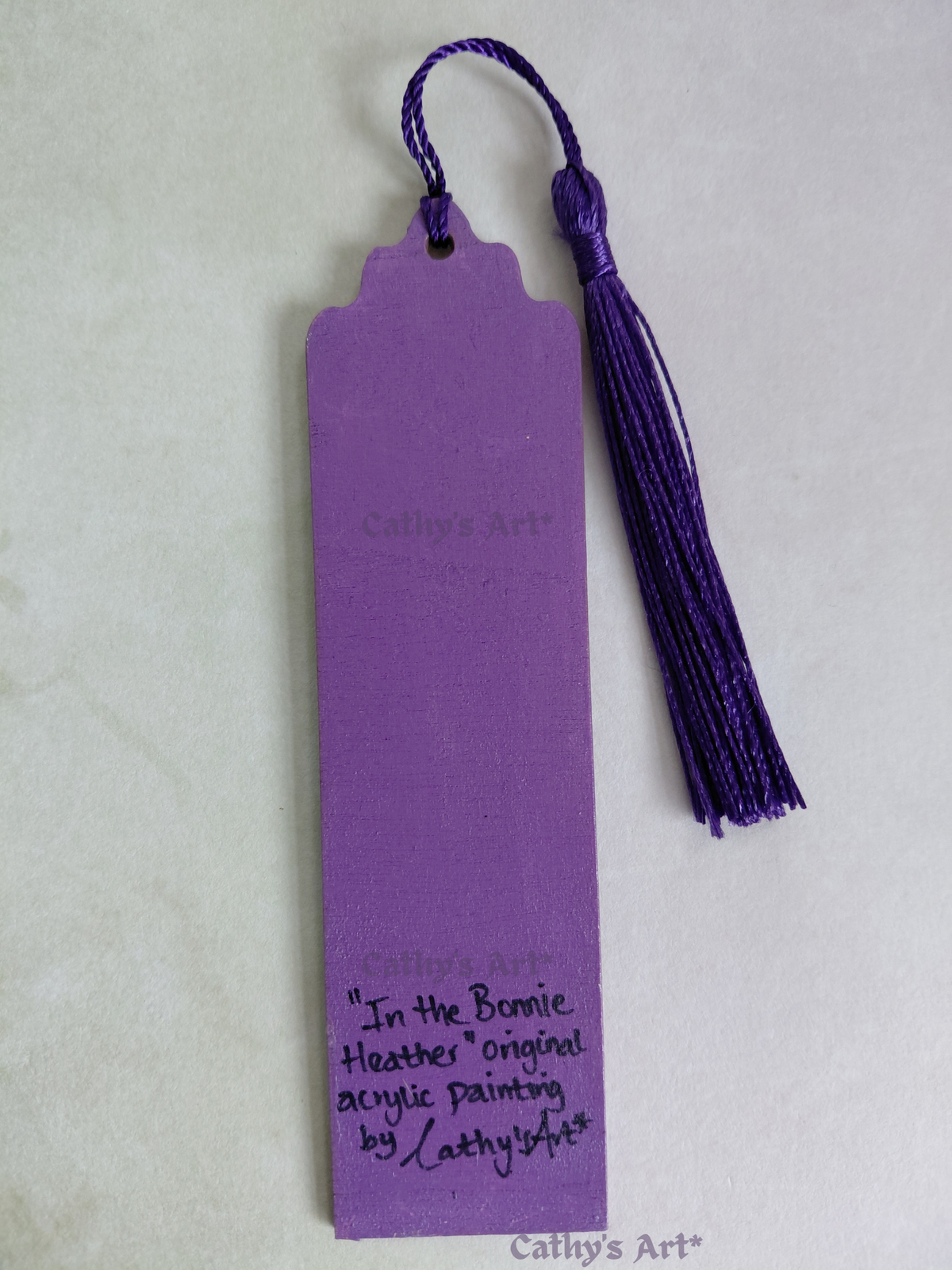 A purple bookmark with a purple tassel, featuring handwritten text, "In the Bonnie Heather" Original acrylic painting by Cathy's Art* on a light background. By Cathy's Art*