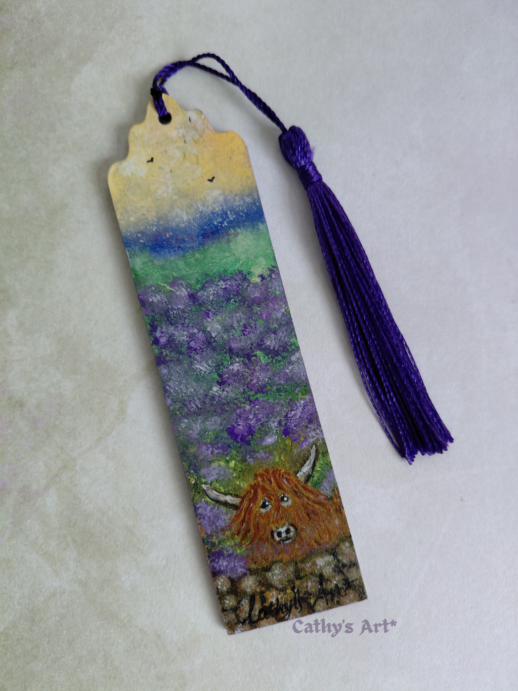A hand-painted wooden bookmark featuring a Highland cow in a lavender field under a blue and yellow sky, complete with a purple tassel, displayed on a textured light  background. By Cathy's Art*