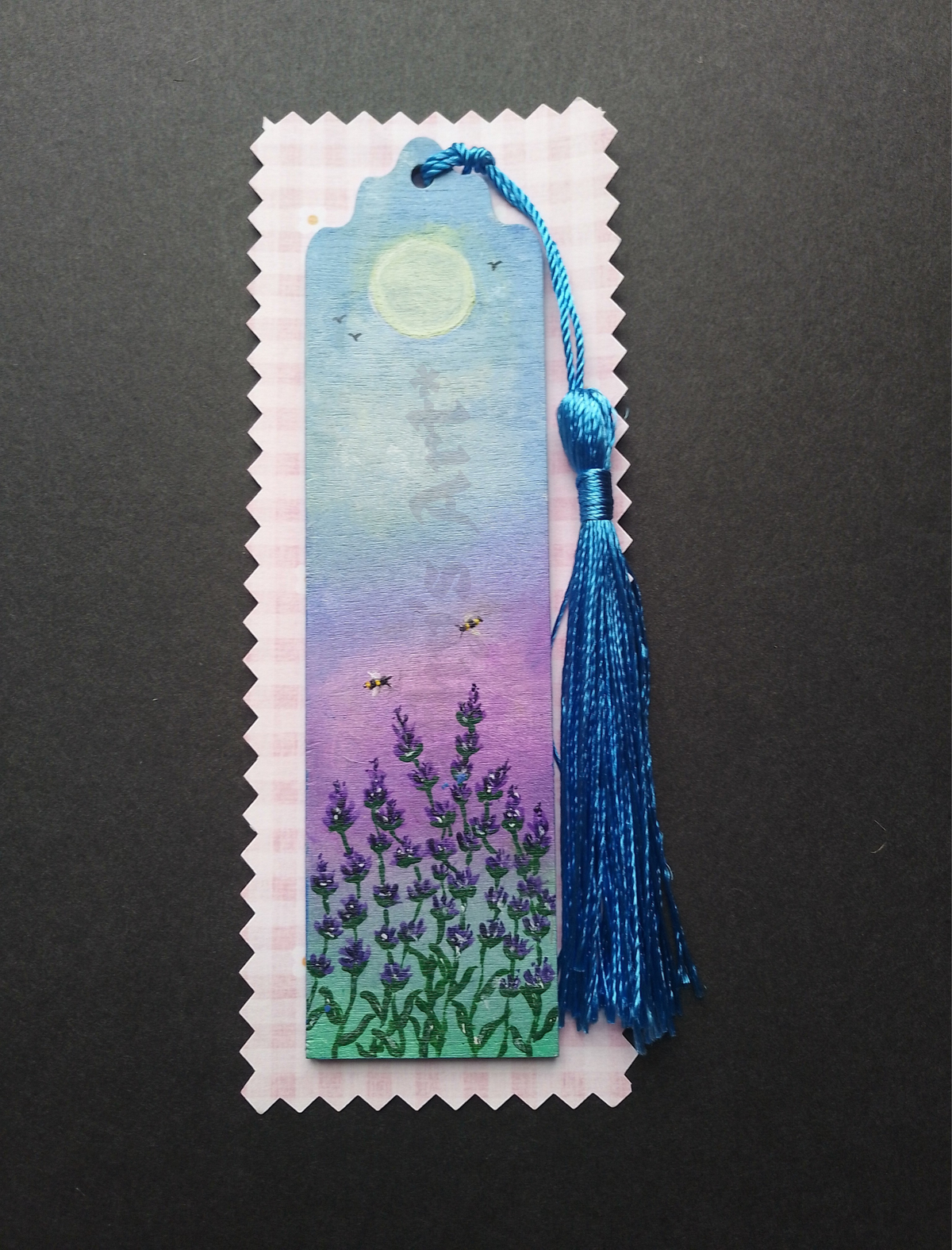 A hand painted bookmark with a blue tassel, featuring a painted lavender field under a yellow sun and purple sky on lilac packaging on a black background. By Cathy's Art*