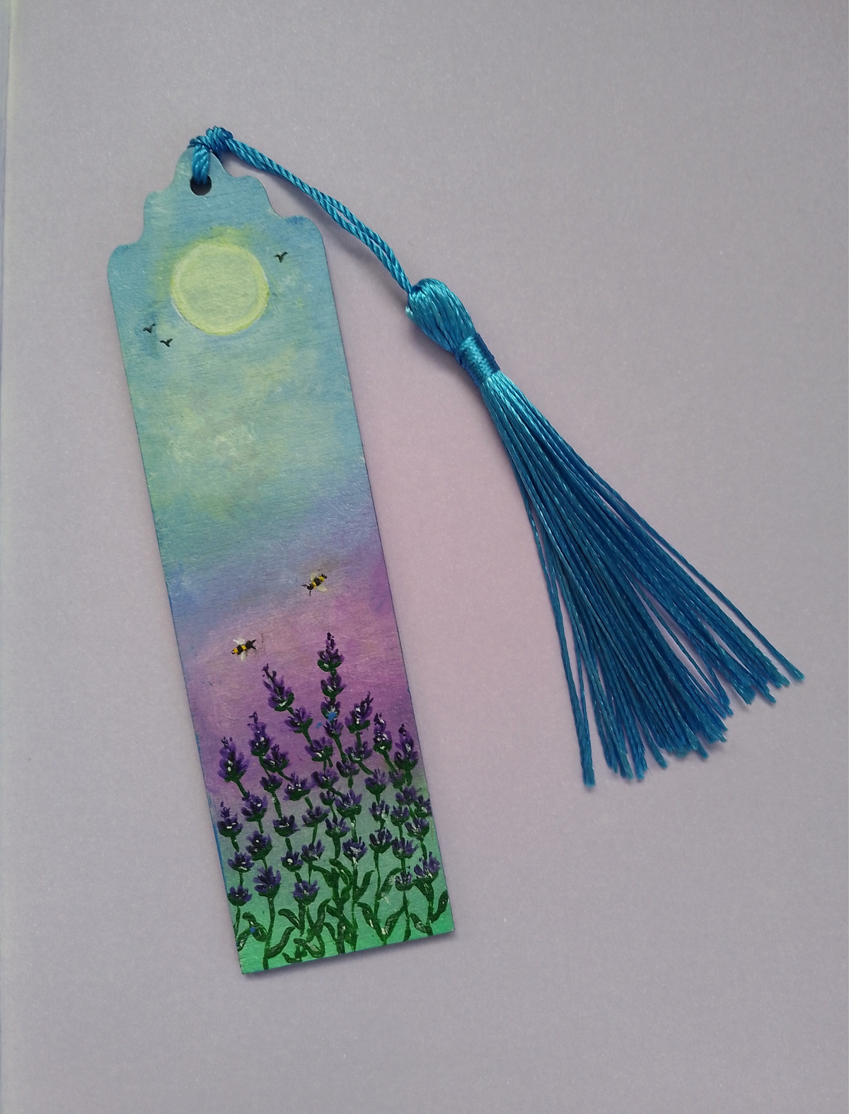 A hand painted bookmark with a blue tassel, featuring a painted lavender field under a yellow sun and purple sky on a lilac background. By Cathy's Art*