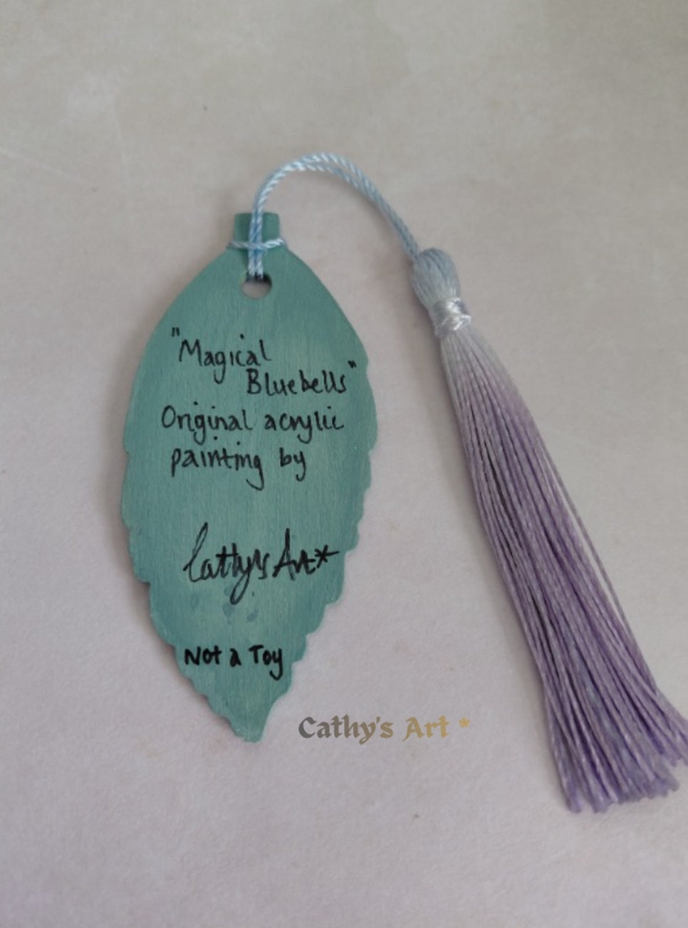 A decorative leaf-shaped bookmark with a tassel, shown on a light background. The bookmark is a muted teal-green colour with handwritten text reading “Magical Bluebells" Original acrylic painting by Cathy's Art” and “Not a Toy. Original hand painted wooden leaf bookmark by Cathy's Art*