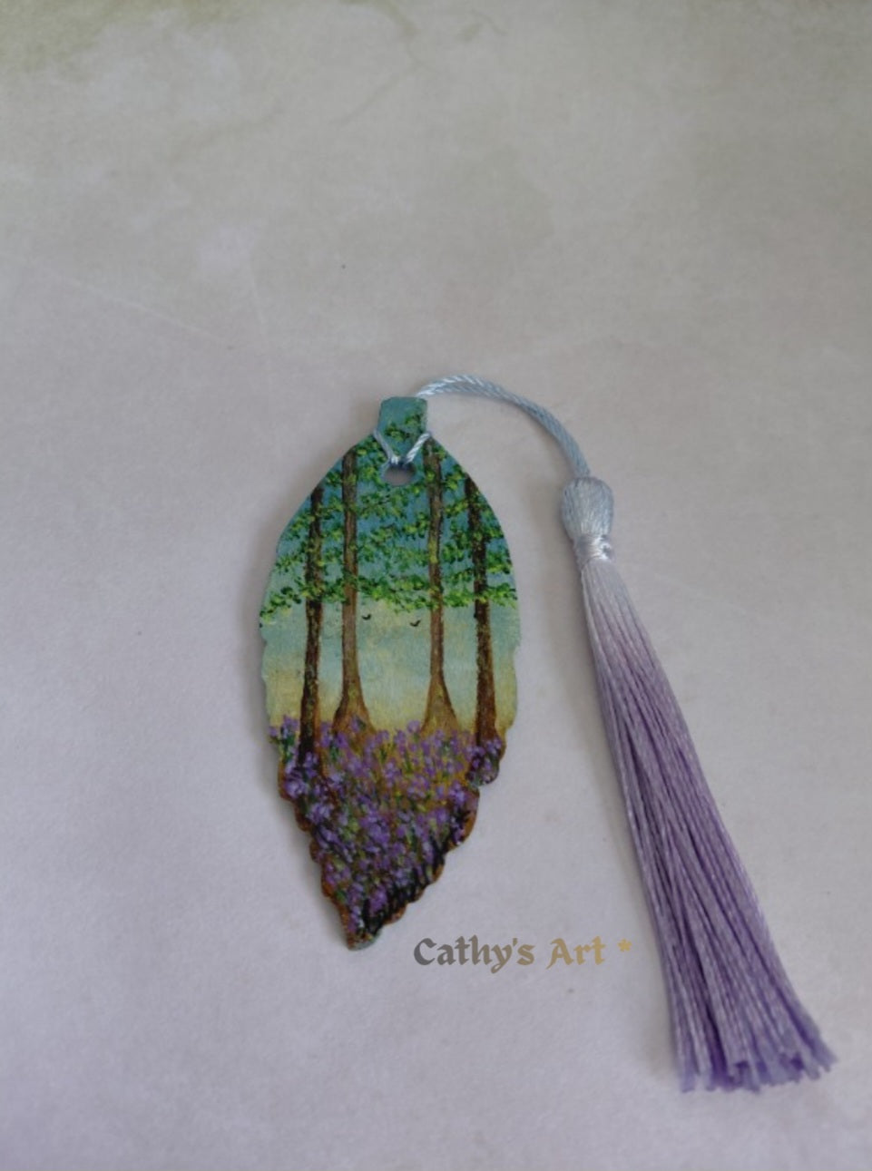 A leaf-shaped bookmark featuring a painted forest scene with green trees and purple bluebell flowers, complete with a blue lavender tassel, displayed on a light background. Original hand painted bookmark by Cathy's Art*