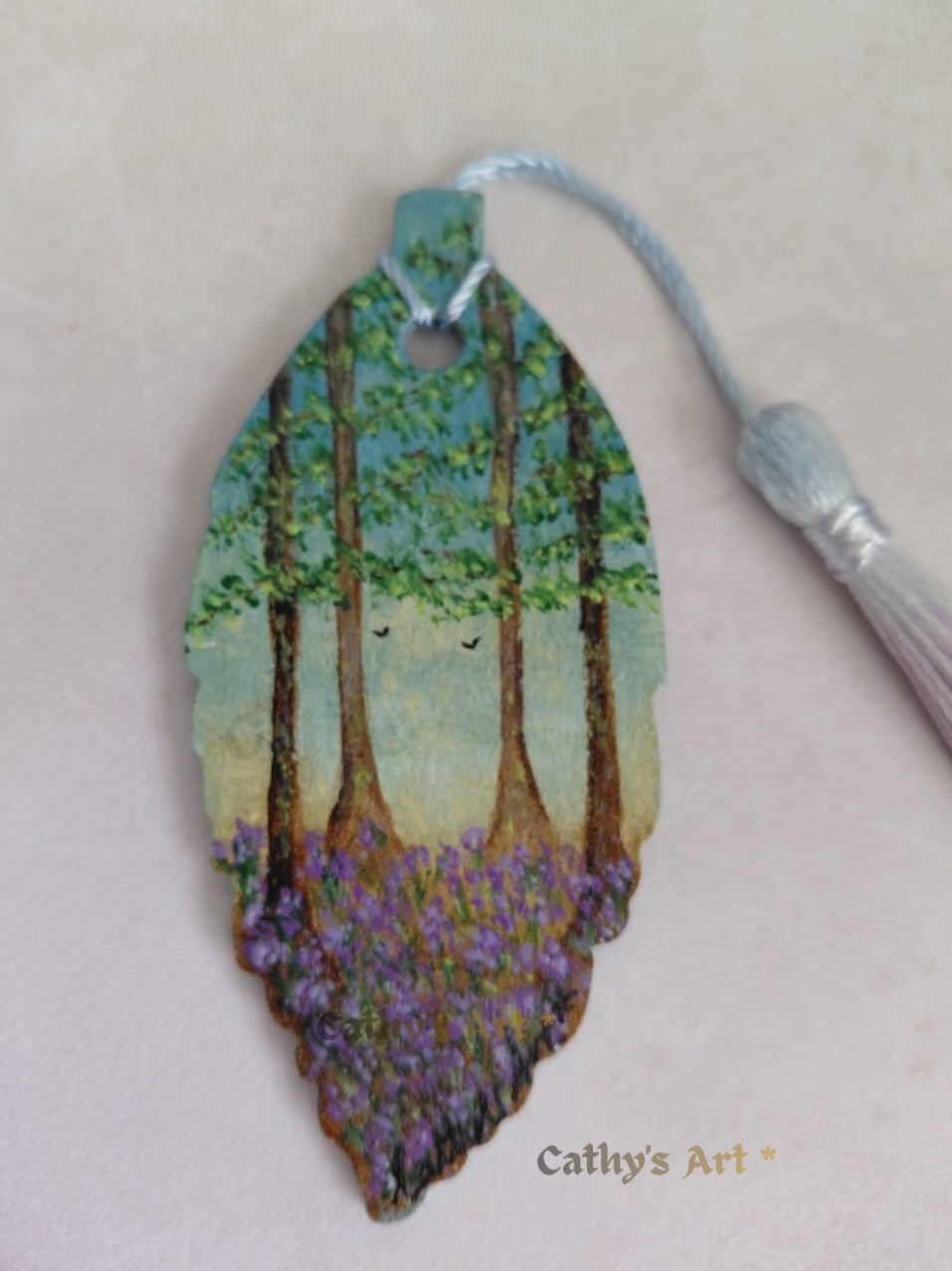 A close up image hand painted leaf-shaped bookmark featuring a forest scene with tall trees, green foliage, and bluebell flowers, birds in the sky, with a light blue tassel attached. The text 'Cathy's Art *' is printed near the bottom. Original hand painted leaf bookmark by Cathy's art*