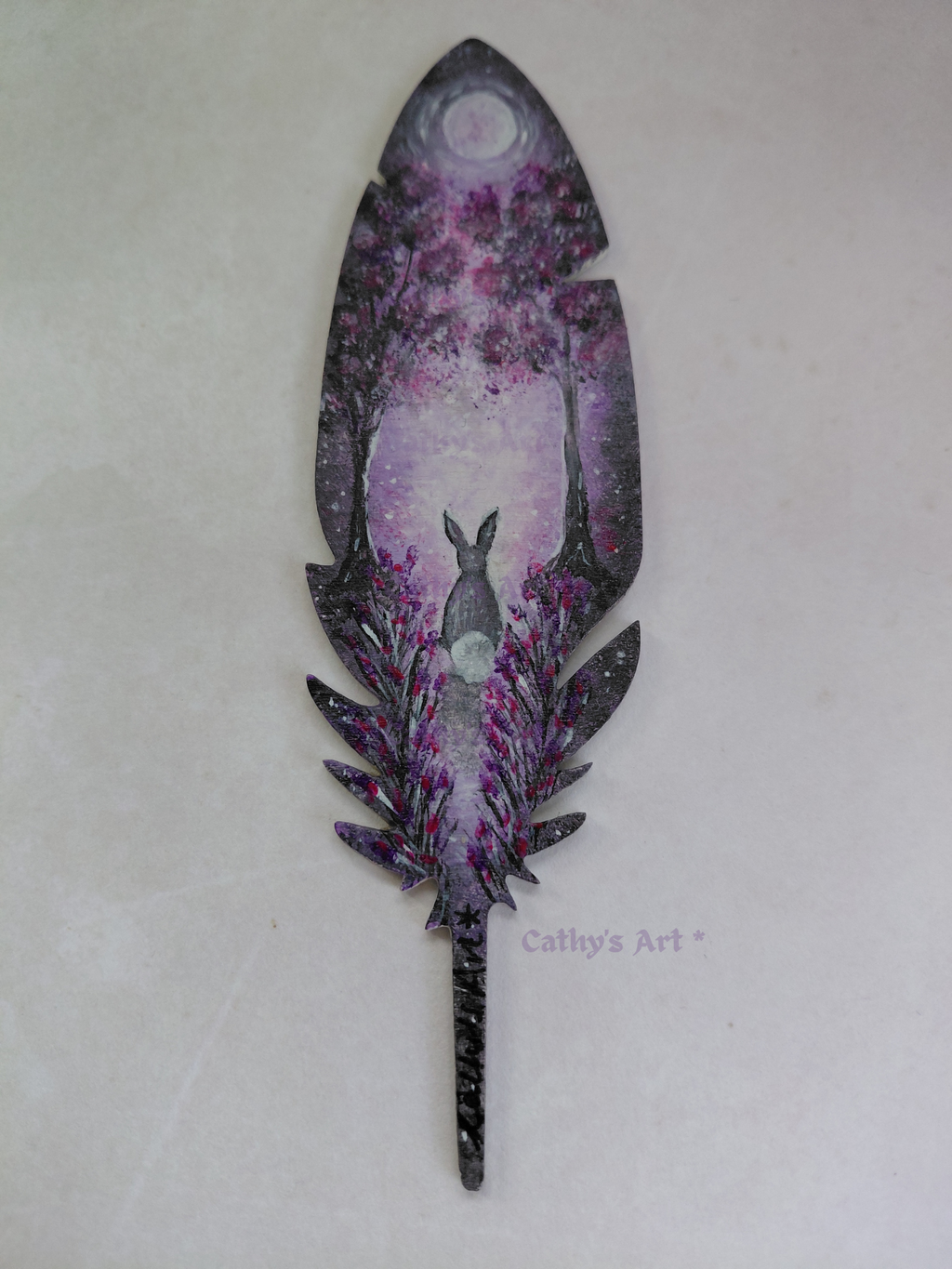 A magical feather shaped bookmark, hand painted with a whimsical purple and violet forest scene. Where a hare sits transfixed by the moon beneath the trees, flowers growing either side of a moonlit pathway. Original acrylic painting on a wooden feather bookmark by Cathy's Art*