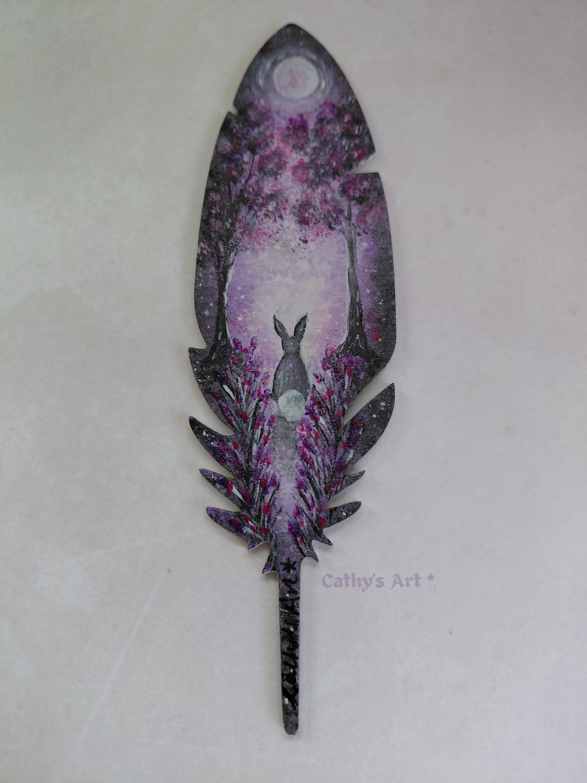 A magical feather shaped bookmark, hand painted with a whimsical purple and violet forest scene. Where a hare sits transfixed by the moon beneath the trees, flowers growing either side of a moonlit pathway. Original acrylic painting on a wooden feather bookmark by Cathy's Art*