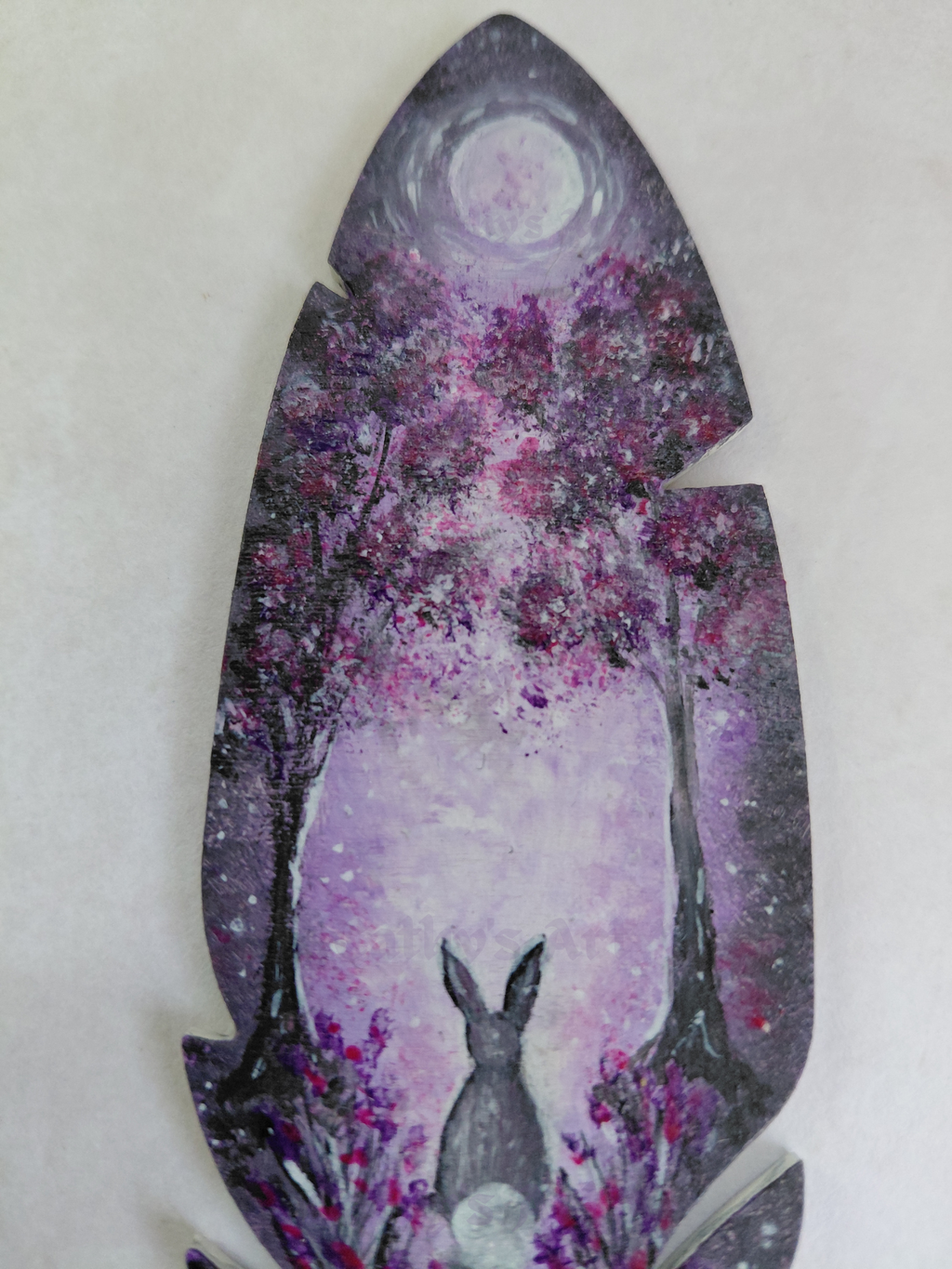 A close up view of the top park of a feather shaped bookmark. Hand painted scene in purple and grey colours. A moon shines through two trees onto a hare that sis below amongst flowers. Original hand painted feather wooden bookmark by Cathy's Art*