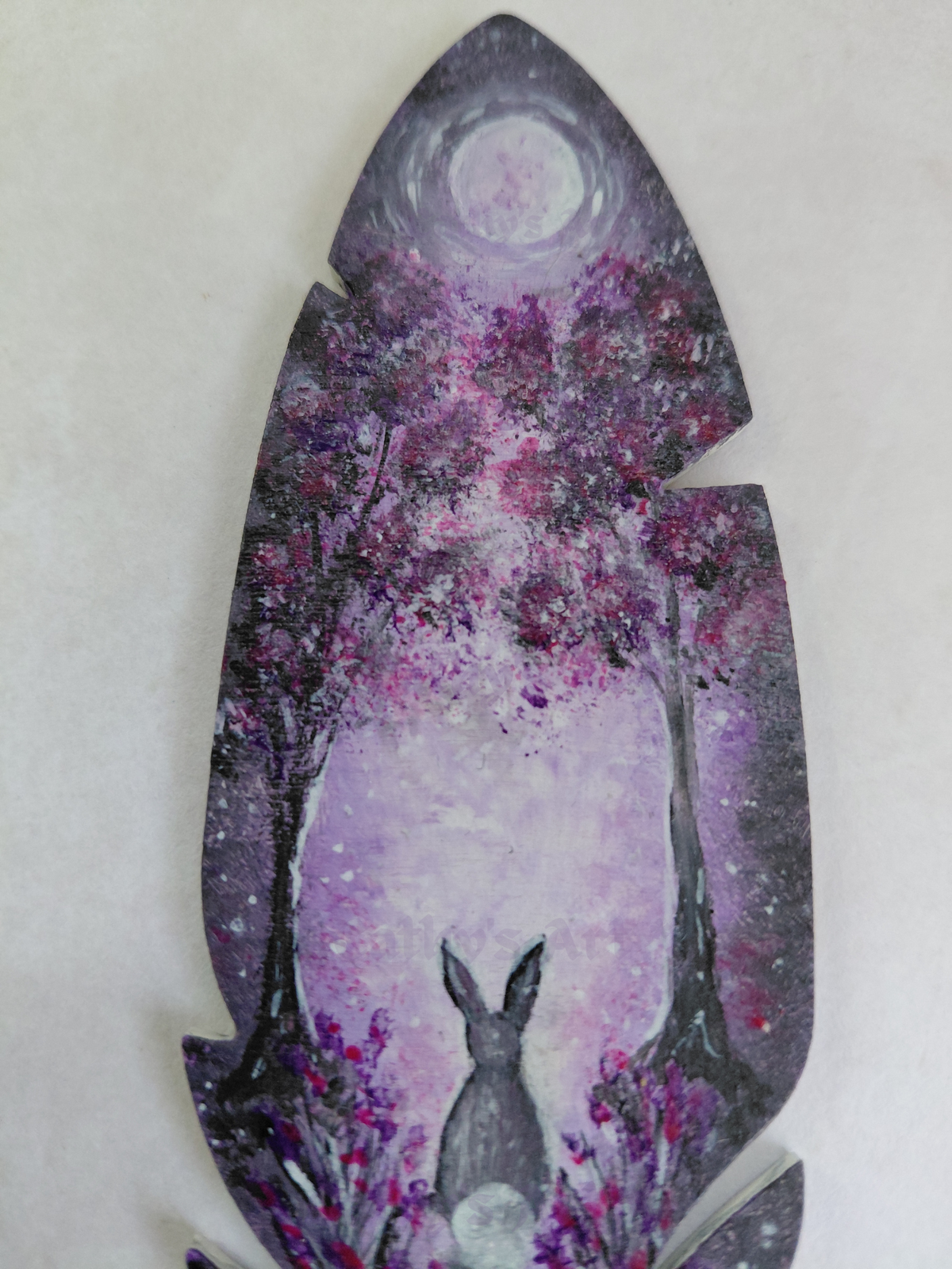 A close up view of the top park of a feather shaped bookmark. Hand painted scene in purple and grey colours. A moon shines through two trees onto a hare that sis below amongst flowers. Original hand painted feather wooden bookmark by Cathy's Art*