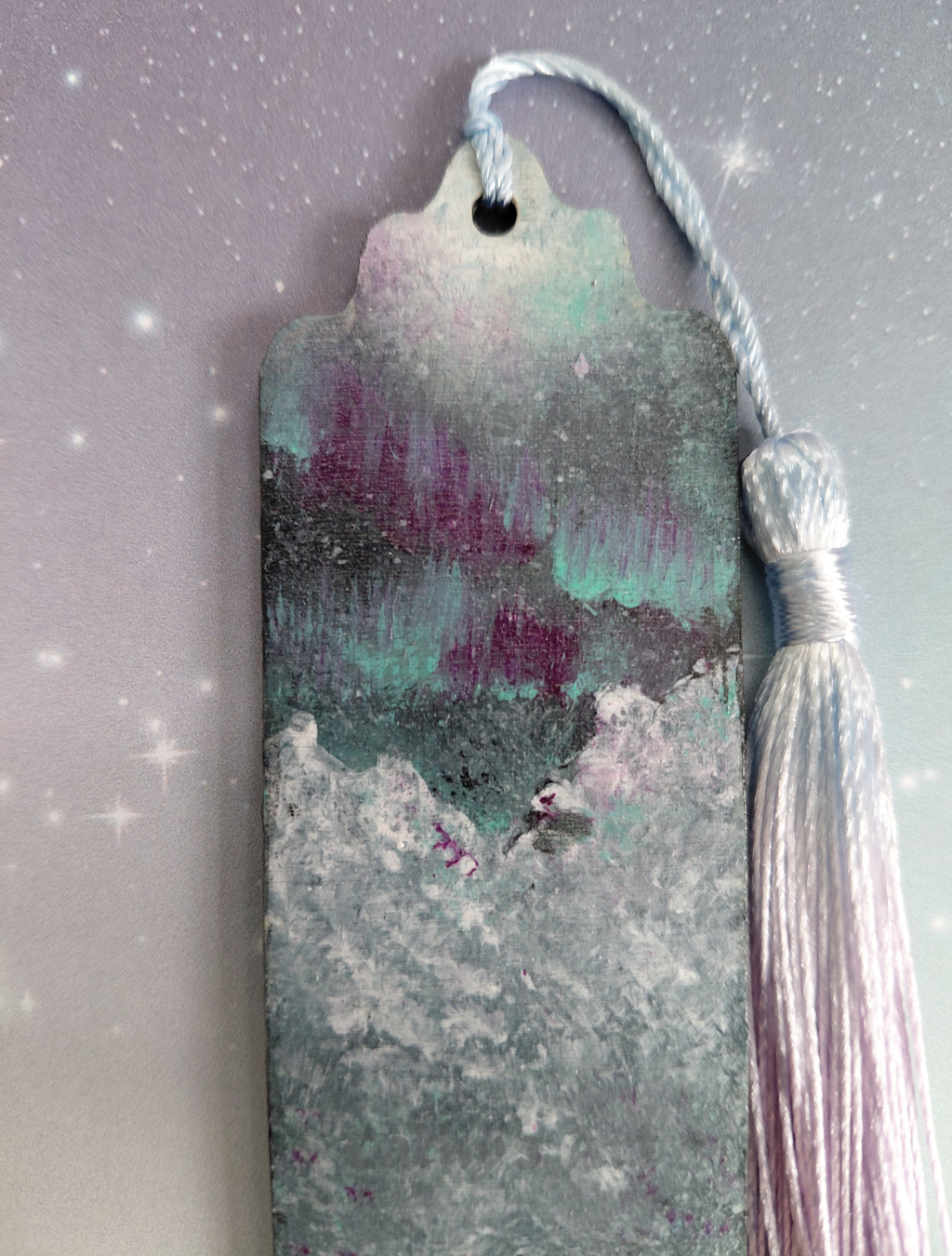 Hand painted top half of a bookmark, featuring the Aurora Borealis sky, white capped snowy mountains, with a blue lilac gradient tassel. Photographed against a purple lilac star background. Original acrylic painted bookmark by Cathy's Art* 
