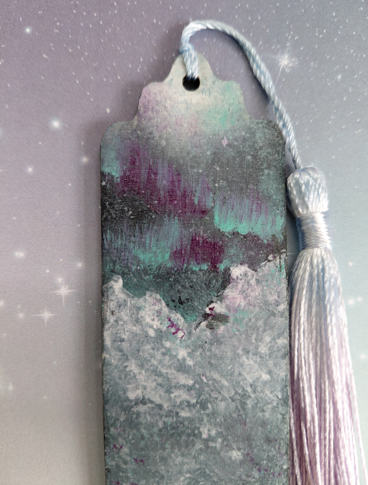 Hand painted top half of a bookmark, featuring the Aurora Borealis sky, white capped snowy mountains, with a blue lilac gradient tassel. Photographed against a purple lilac star background. Original acrylic painted bookmark by Cathy's Art* 