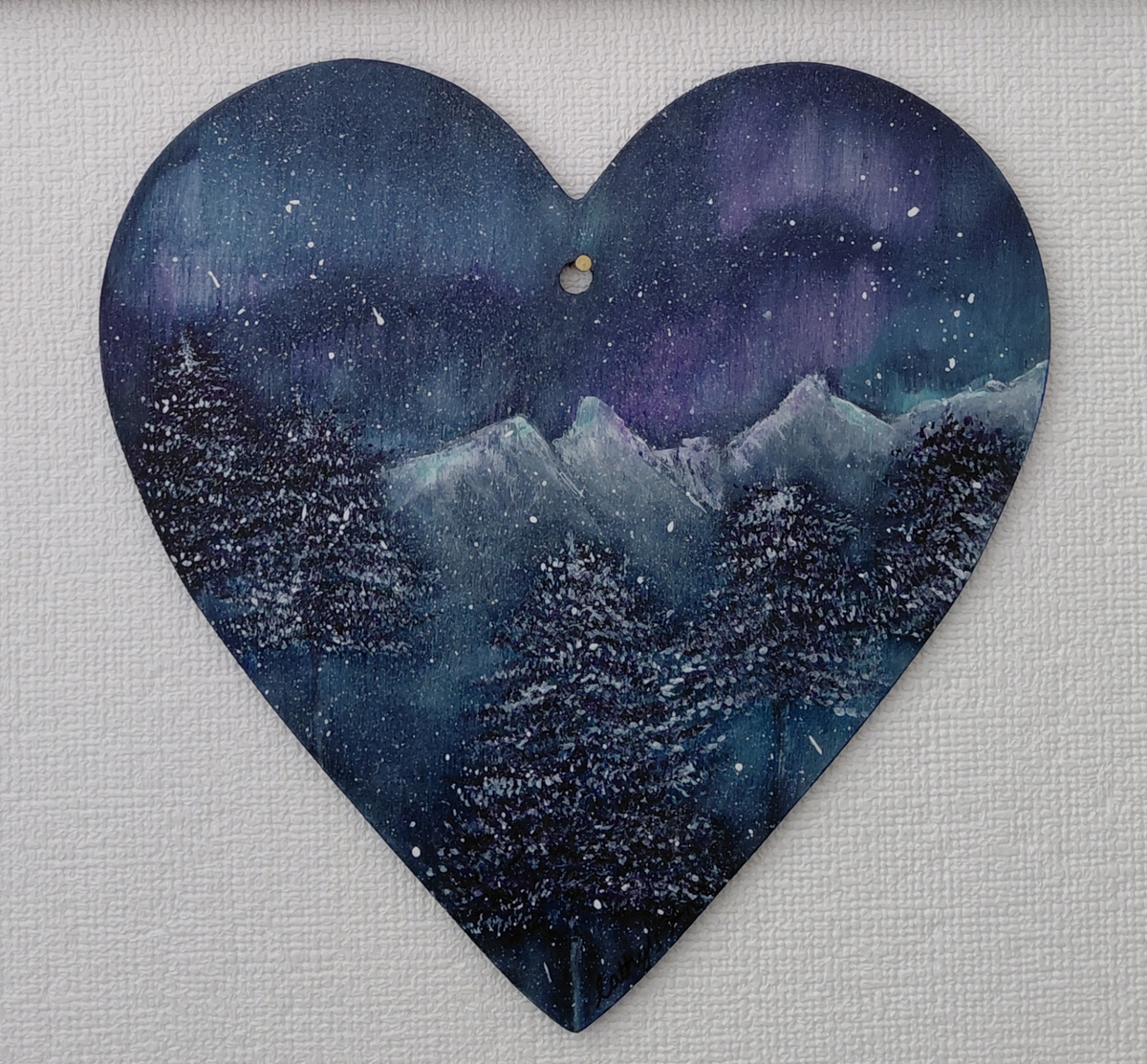 A winter scene on a heart shape, with dark fir trees, snow capped mountains, featuring the Aurora Borealis in the sky in colours of purples and turquoise blues on a dark grey background. Original acrylic painting on wooden heart by Cathy's Art*