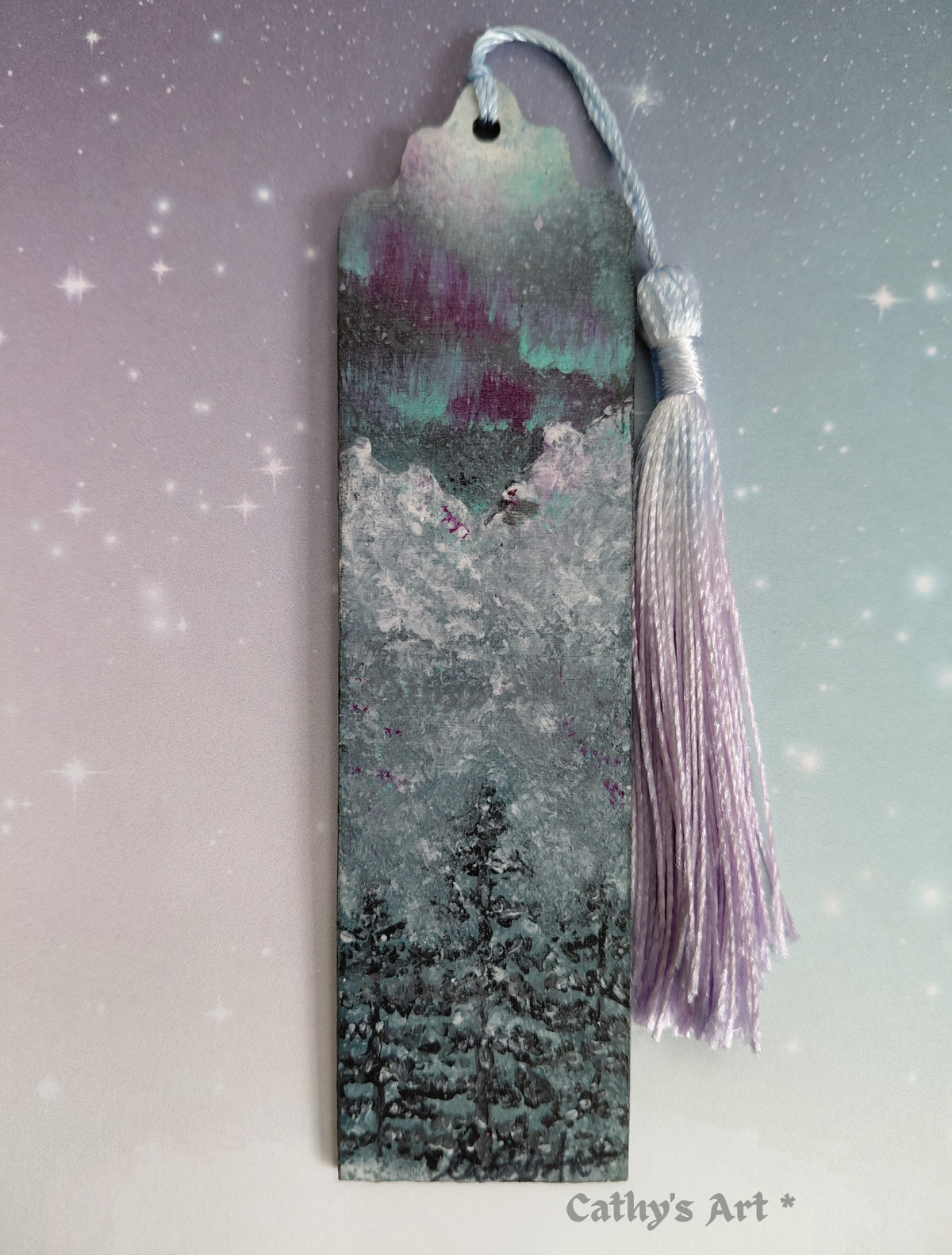 A hand painted bookmark with a tassel, featuring a painted winter forest scene with snowy trees and a purple aurora sky. The bookmark is set against a soft gradient background with star-like sparkles, and the text 'Cathy's Art *' appears at the bottom right. Original hand painted bookmark by Cathy's Art*