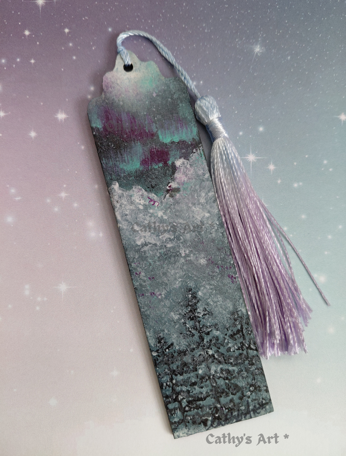 A hand painted bookmark with a tassel, featuring a painted winter forest scene with snowy trees and a purple aurora sky. The bookmark is set against a soft gradient background with star-like sparkles, and the text 'Cathy's Art *' appears at the bottom right. Original hand painted bookmark by Cathy's Art*