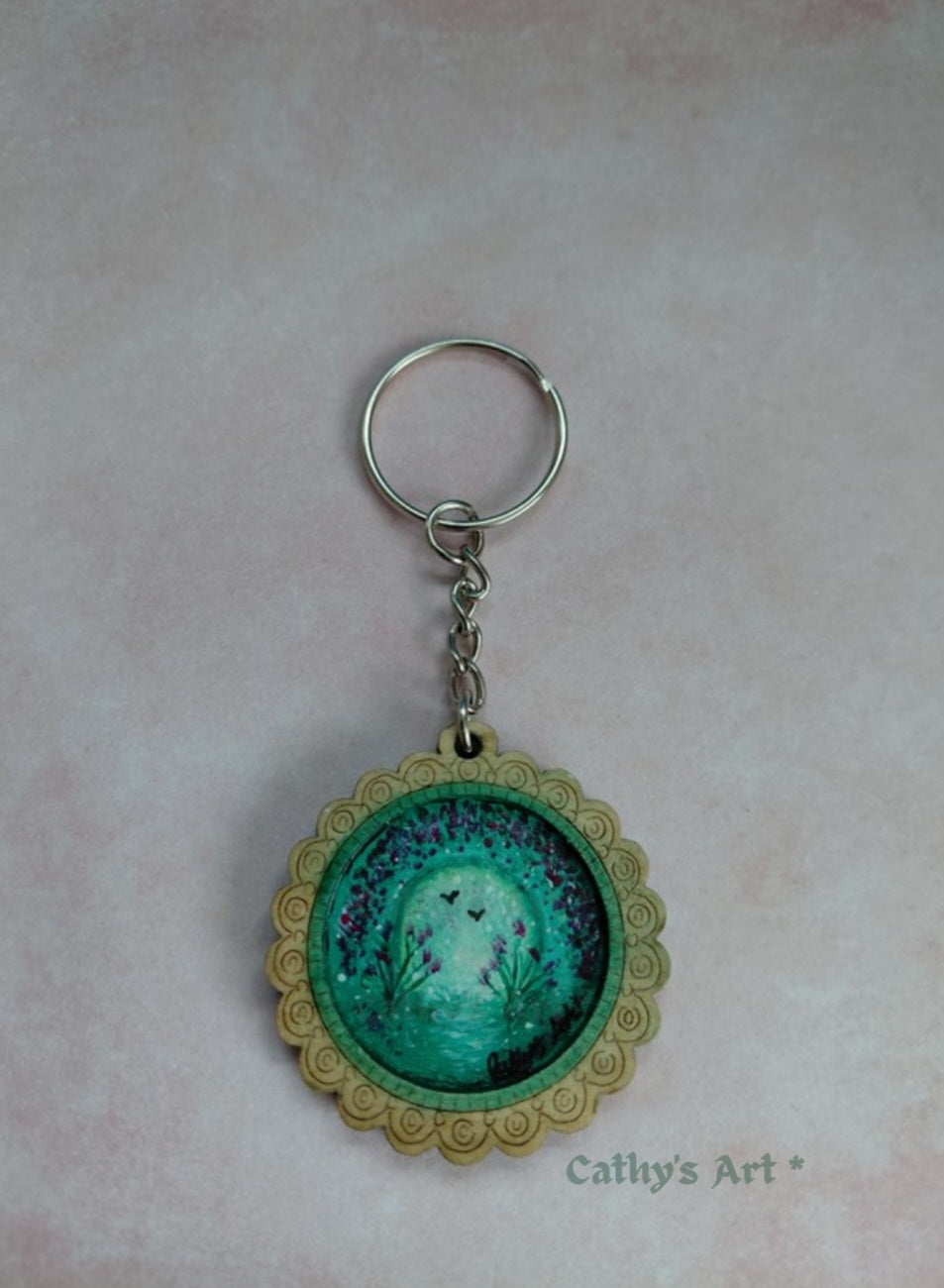 A circular keychain featuring a fantasy art painting of a glowing green and violet mystical land, framed by scalloped border, with a silver metal key ring and chain, set against a soft pink textured background. Original hand painted keyring by Cathy's Art*
