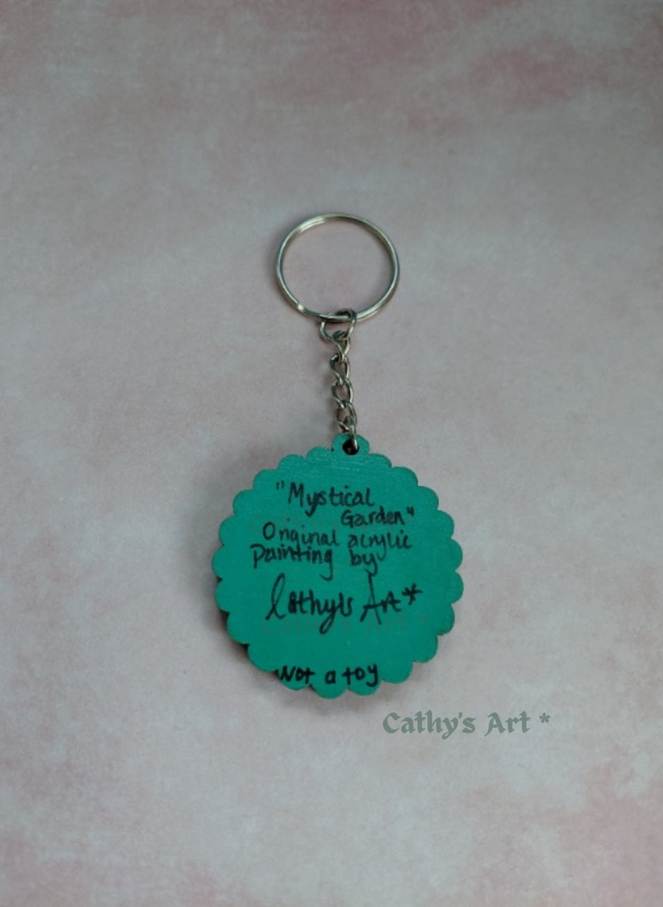 A teal scalloped acrylic keychain with a silver metal split ring and chain, featuring handwritten text that reads 'Mystical Garden Original acrylic painting by Cathly's Art*', displayed on a light pink textured background.