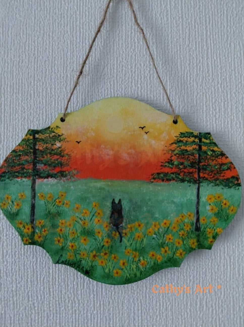 A scalloped wooden hanging plaque featuring a hand-painted landscape of a black cat in a field of yellow flowers at sunset, featuring two trees and birds flying in the sky. Hung on a white textured wall with jute string. Original acrylic painting on a wooden plaque by Cathy's Art*