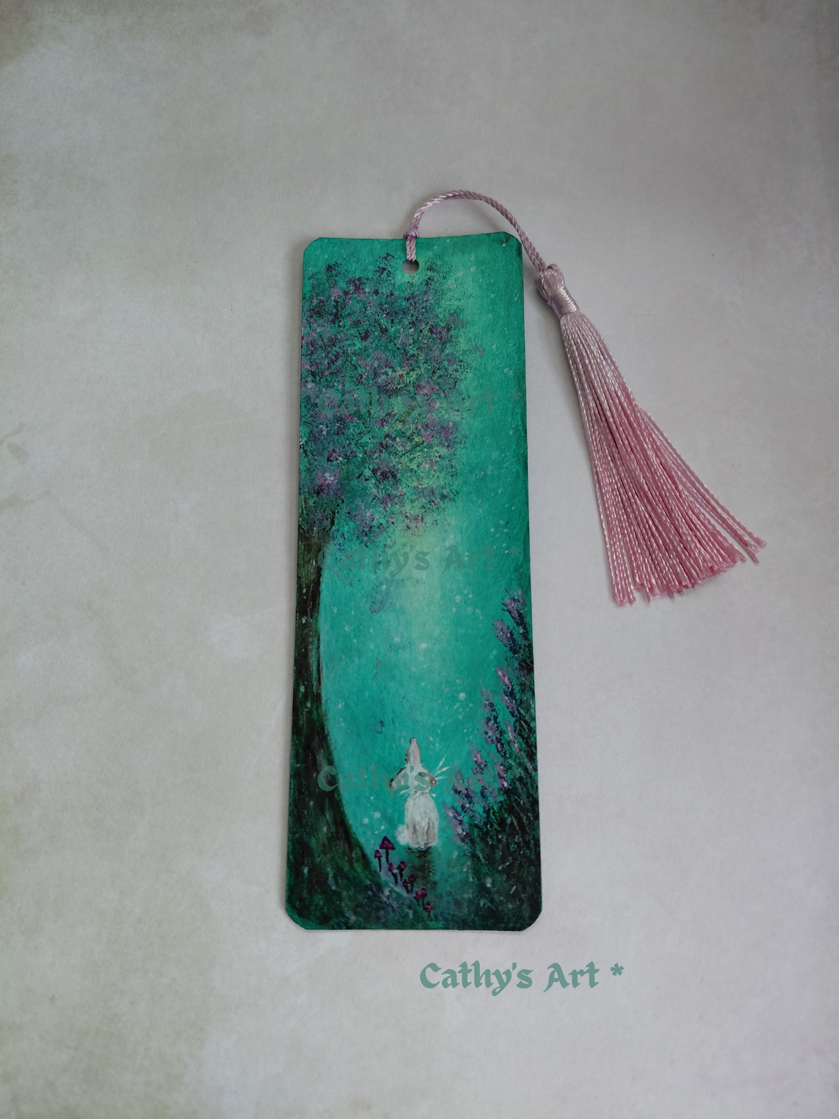 A rectangular bookmark featuring a hand painted whimsical scene of a white rabbit under a glowing pink and green tree, with a pink tassel hanging from the top. The bookmark is displayed on a textured light background with the text 'Cathy's Art  *' printed below. Original hand painted bookmark by Cathy's Art*