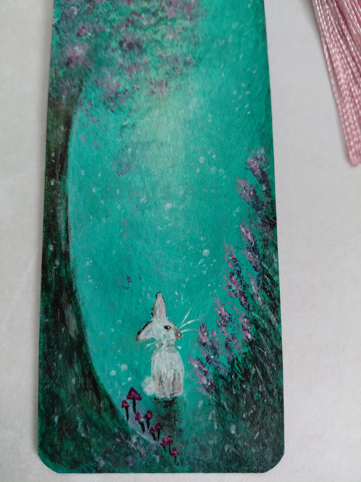 A close - up hand-painted bookmark featuring a whimsical forest scene with a white rabbit, pink flowers, mushrooms and under a turquoise green sky. On a light coloured background. Original acrylic hand painted bookmark by Cathy's Art*