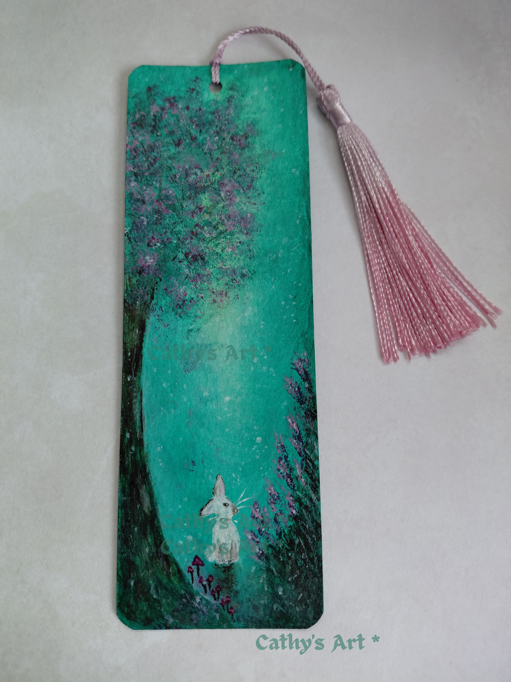 A rectangular hand-painted bookmark featuring a whimsical forest scene with a white rabbit, pink flowers, mushrooms and under a turquoise green sky, complete with a pink tassel. Original acrylic hand painted bookmark by Cathy's Art*