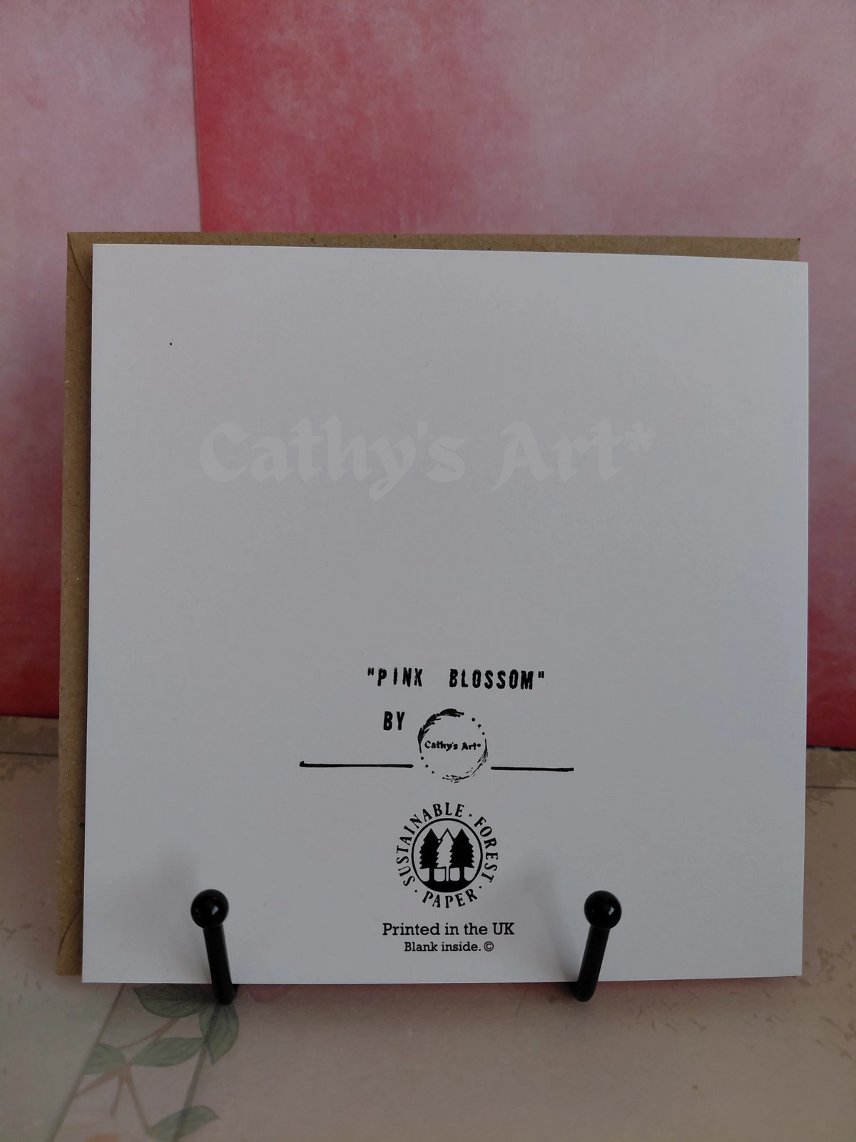A white art printed greeting card with black text reading "Pink Blossom" by Cathy's Art*, displayed on a kraft paper backing with black stand clips against a pink background. Original printed greeting card by Cathy's Art