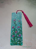 A rectangular turquoise green hand painted bookmark with a pink tassel, decorated with pink and white daises with and green stems, and two bees. Photographed on a light background. Original acrylic painting on watercolour paper bookmark by Cathy's Art*